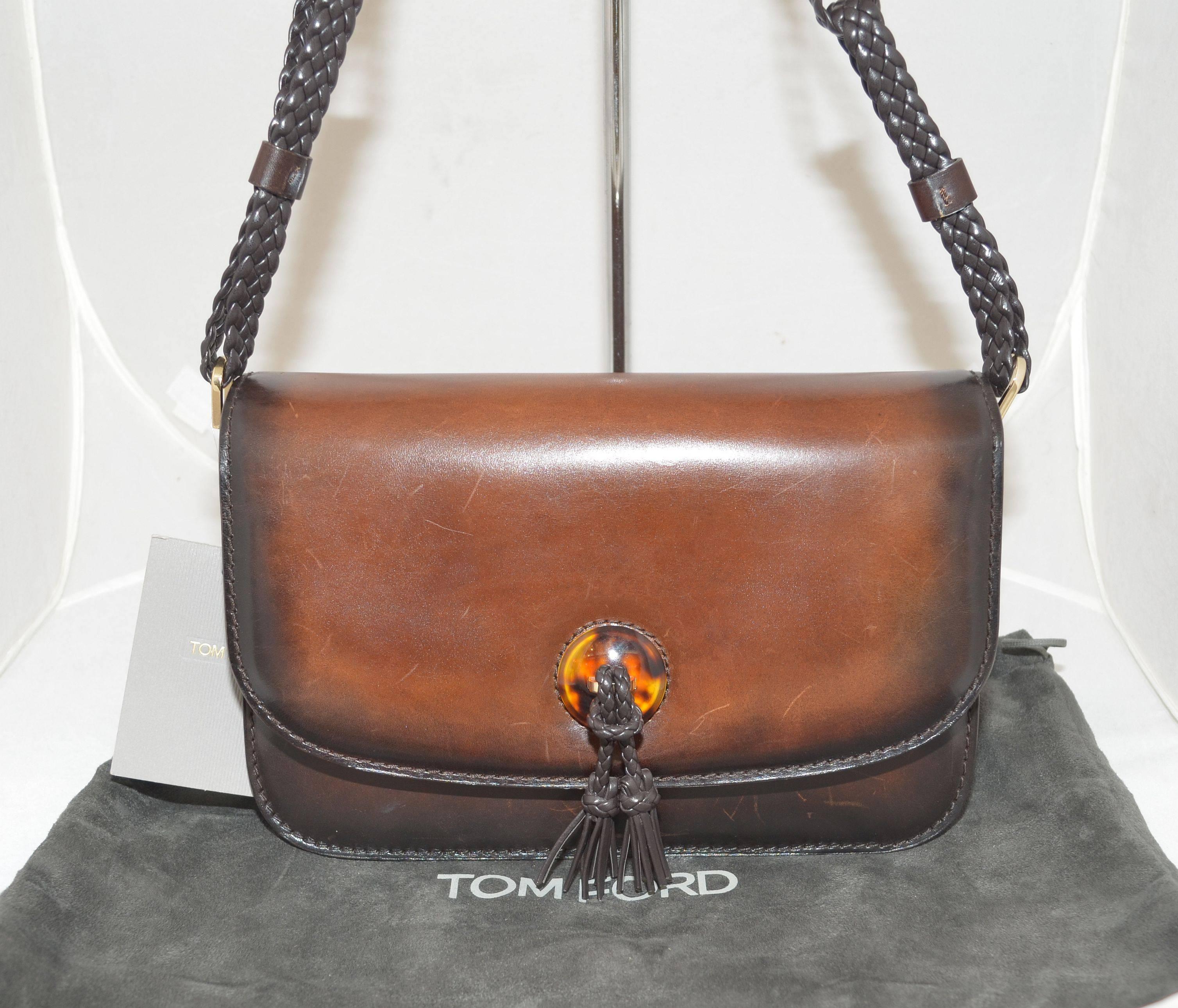 Tom Ford bag features a braided leather shoulder strap, magnetic snap closure, tassel applique at the front center, and a tortoise trim around the base of the bag. Interior has three compartments; 2 open compartments and a center zipper compartment.