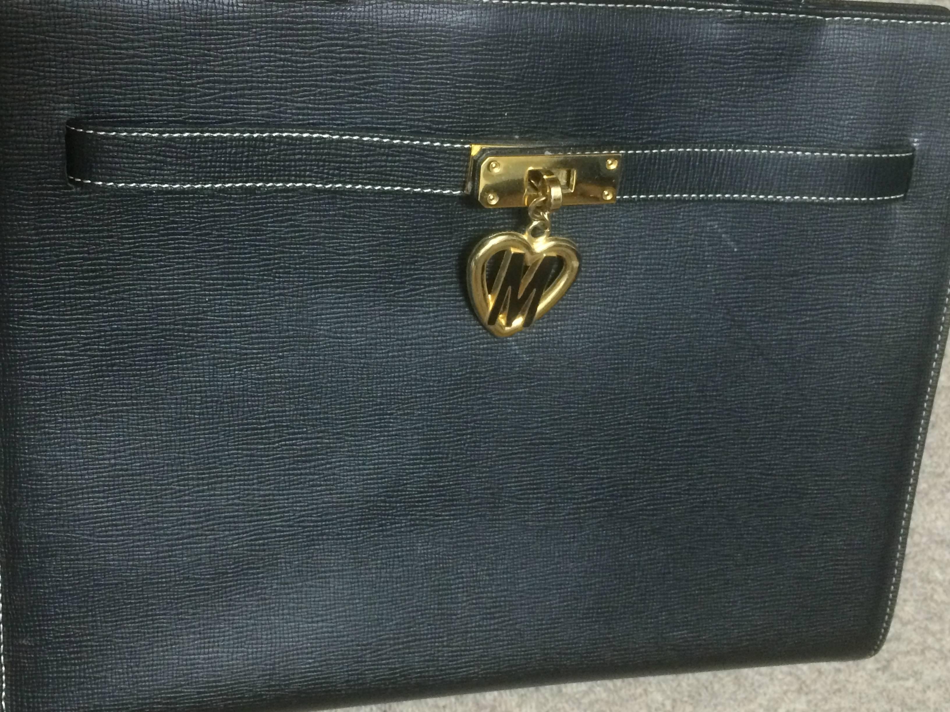 1990s. Vintage MOSCHINO black leather tote bag in Kelly purse style with iconic M and heart charm.

This classic kelly style tote bag from MOSCHINO would never go out of style.
Featuring th 