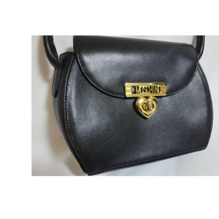 1990s. Vintage MOSCHINO black leather handbag, oval shape purse with a golden logo closure with heart shape charm.

Introducing a vintage black leather cute oval shape handbag from MOSCHINO, approx in the mid to late 90's.
Featuring a gold tone logo