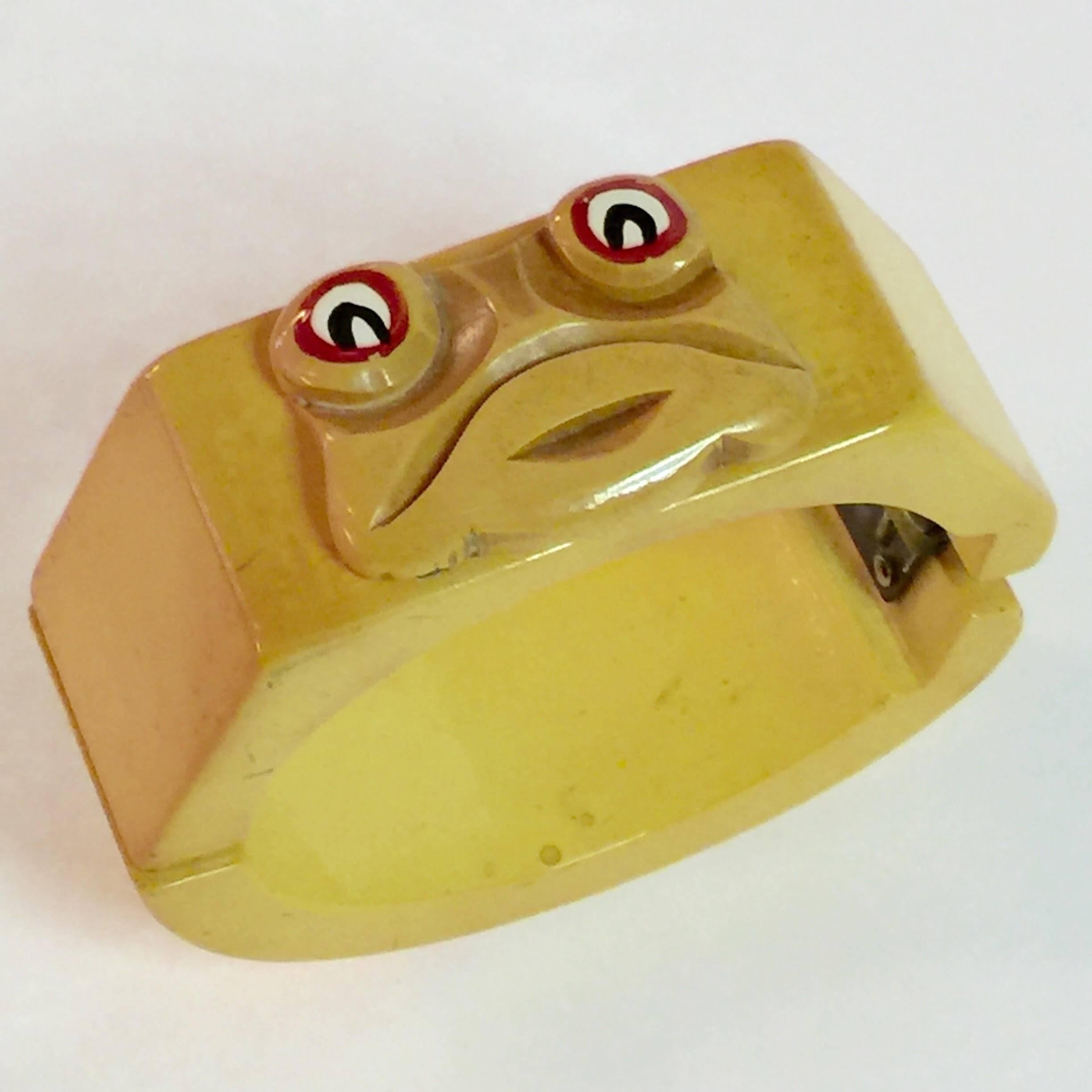 A unique piece of bakelite artistry, this very special hinged bracelet in cream is quite simple, but has an applied whimsical face of bug-eyed frog face on its  top surface. Classic figural bakelite hinge bracelets frequently feature a thematic