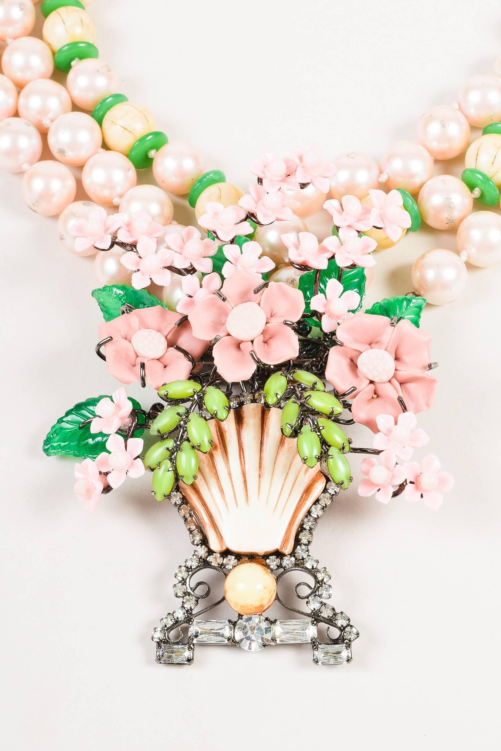 Large pale pink faux pearl strands are details with green and wooden beads. Flower basket pedant has a tan seashell and bead at center with rhinestone trim. Pink flowers at center have green leaves below and fanning the top. Hook closure in