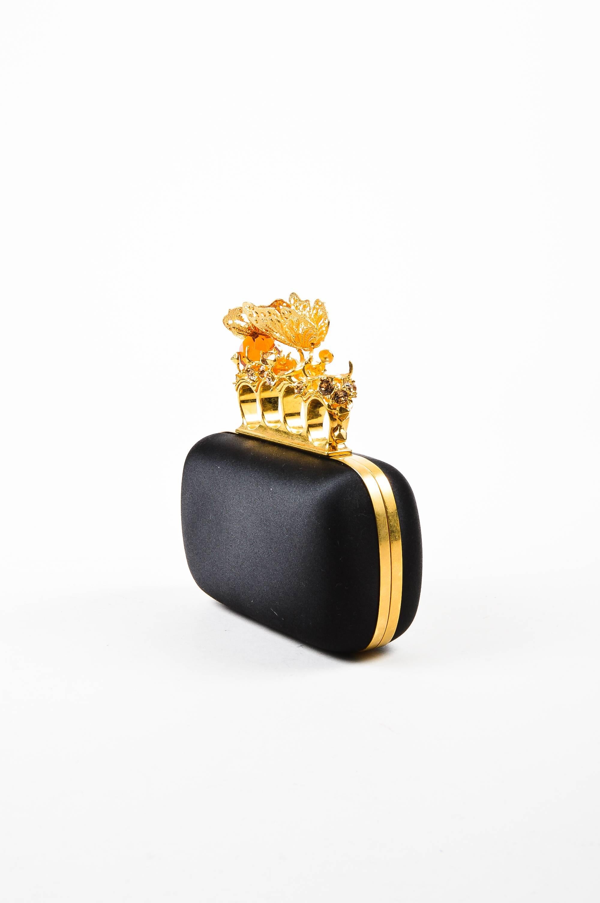 Comes in box with dust bag. Striking black satin clutch is trimmed with gold-tone metal. Push lock closure at top. Knuckle carry top is embellished with gorgeous crystal stones and metal butterflies. Spiked stud details and small skull hiding