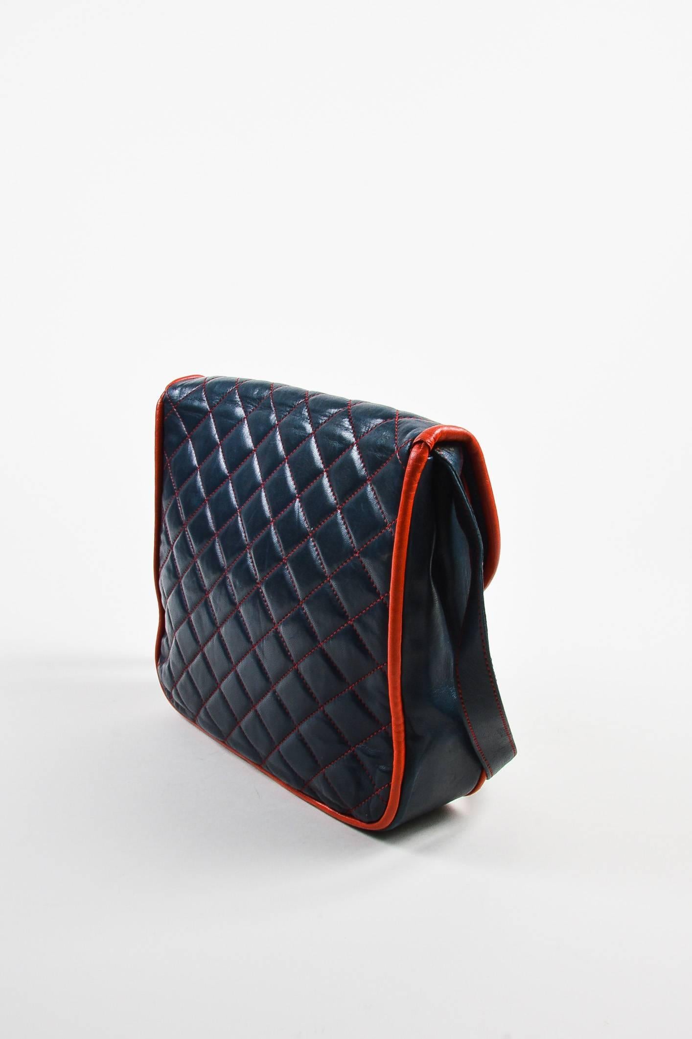 Versatile bag constructed of navy blue leather. Red accents at piping and quilted topstitching. Flap top with snap closure. Flat leather shoulder strap. Spacious interior lined with ribbed textile. Circa 1980s.

Interior Features: Zip
