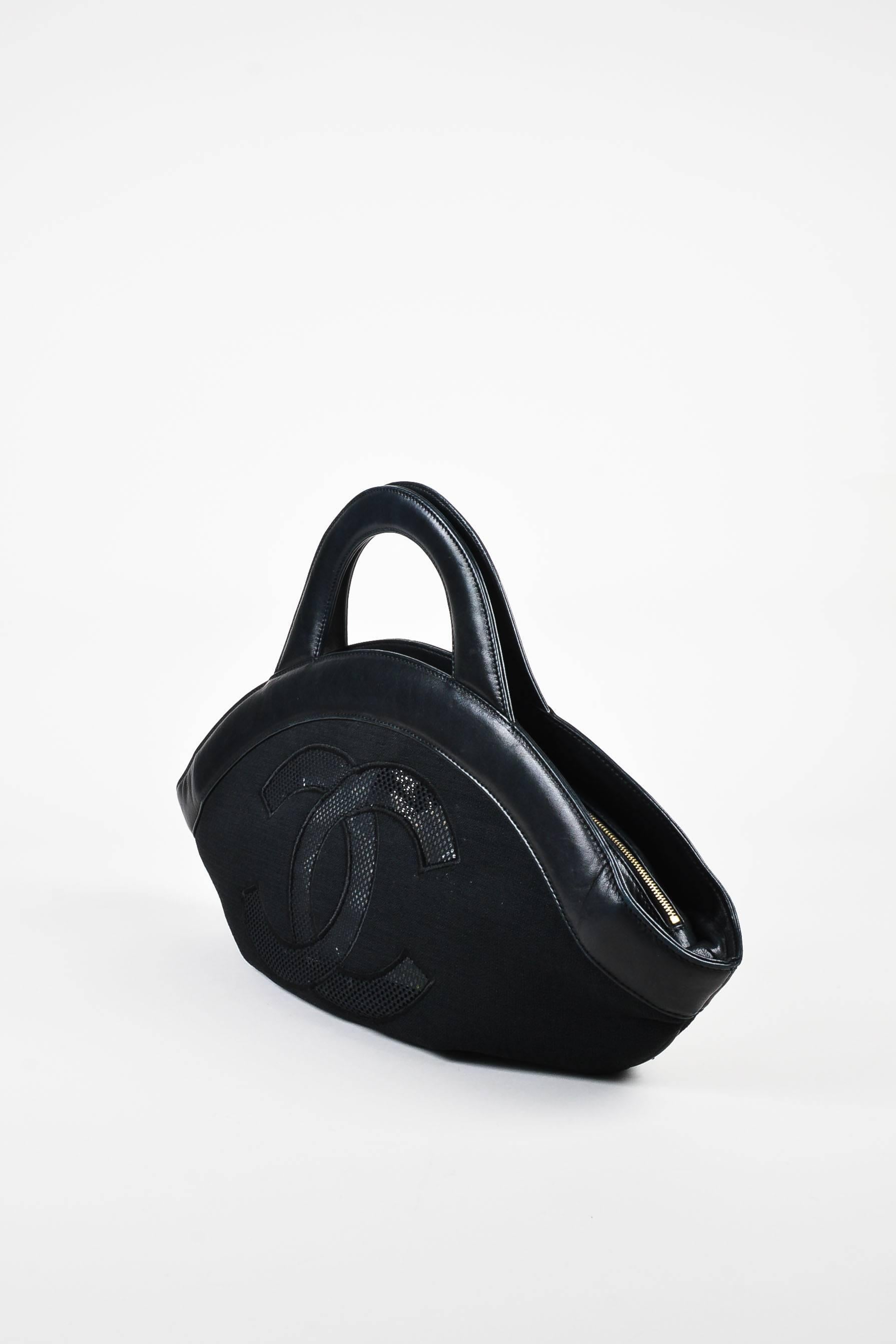 Black canvas handle bag from Chanel circa 2002-2003 has a black leather trim. The front of the bag features a mesh embroidered camellia flower, while the back features a mesh embroidered 'CC' logo. Two handles for wear. Top zipper for closure with a