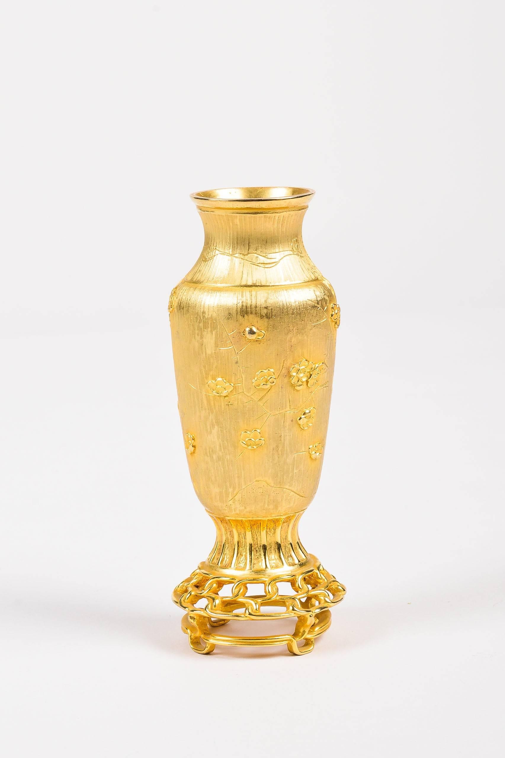 A rare piece by Judith Leiber. This Asian-inspired urn shaped bud vase is constructed of gold-tone metal and depicts images of cherry blossoms and a bird. Etched tree trunk, branches, and grass in background. Vase sits atop an interwoven footed