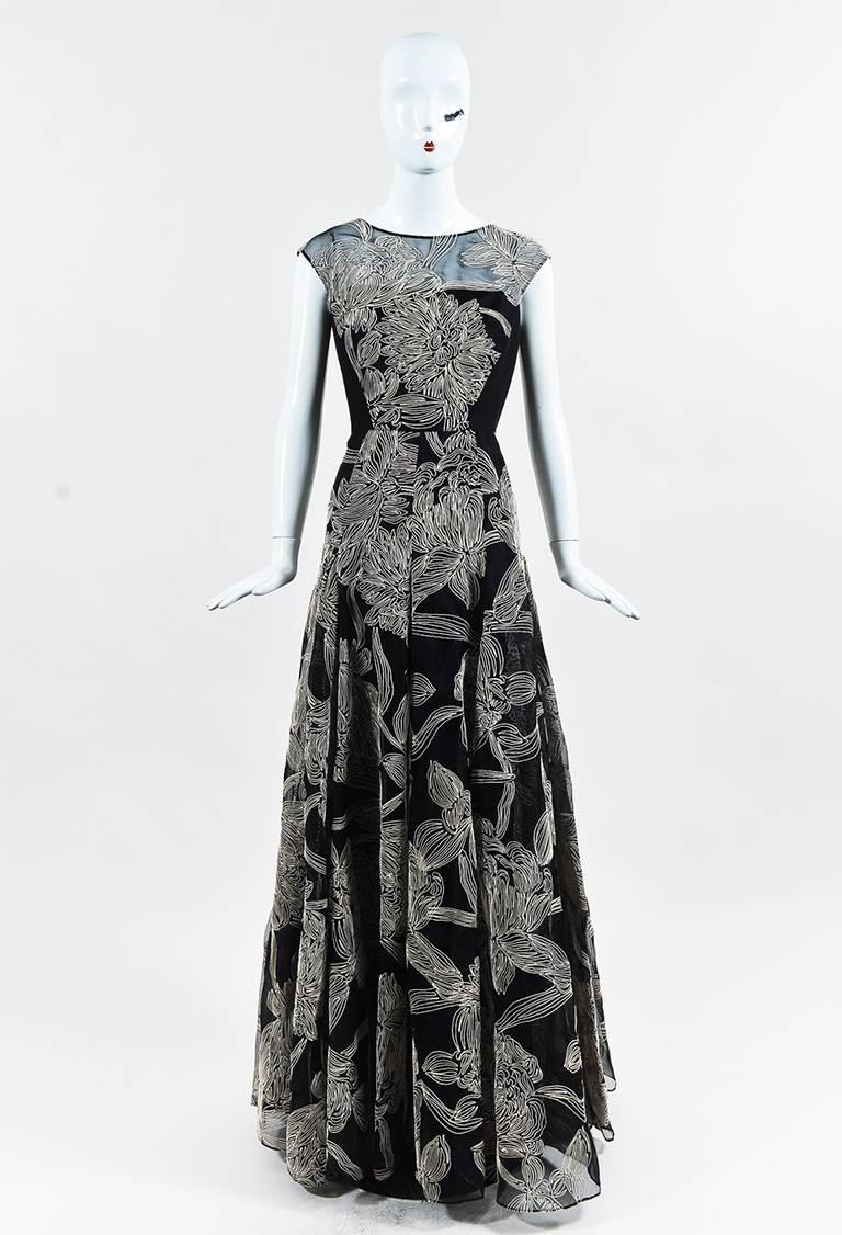 Black silk sleeveless princess cut gown from Carmen Marc Valvo Couture features cream colored floral embroidery. Boat neckline. Sheer paneling on the neckline. Full, pleated skirt with a tulle underlay for additional volume. Back invisible zipper