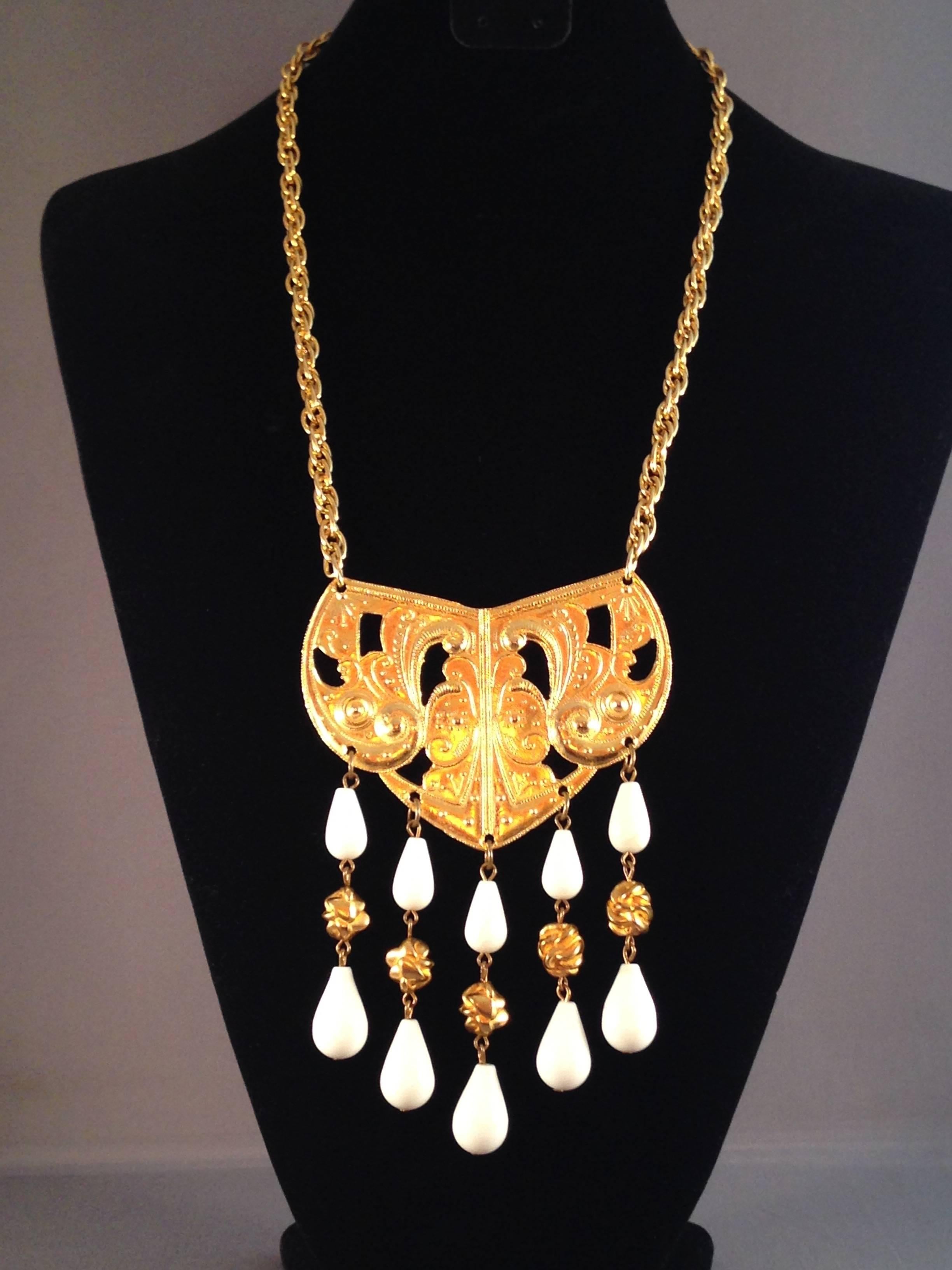 This is a 1970s Kenneth Jay Lane gold tone pendant necklace. The pendant measures 4 1/2" long in total x 3 1/4" wide. It has five strands that hang from the pendant made up of white lucite and gold tone beads. The chain measures 19"