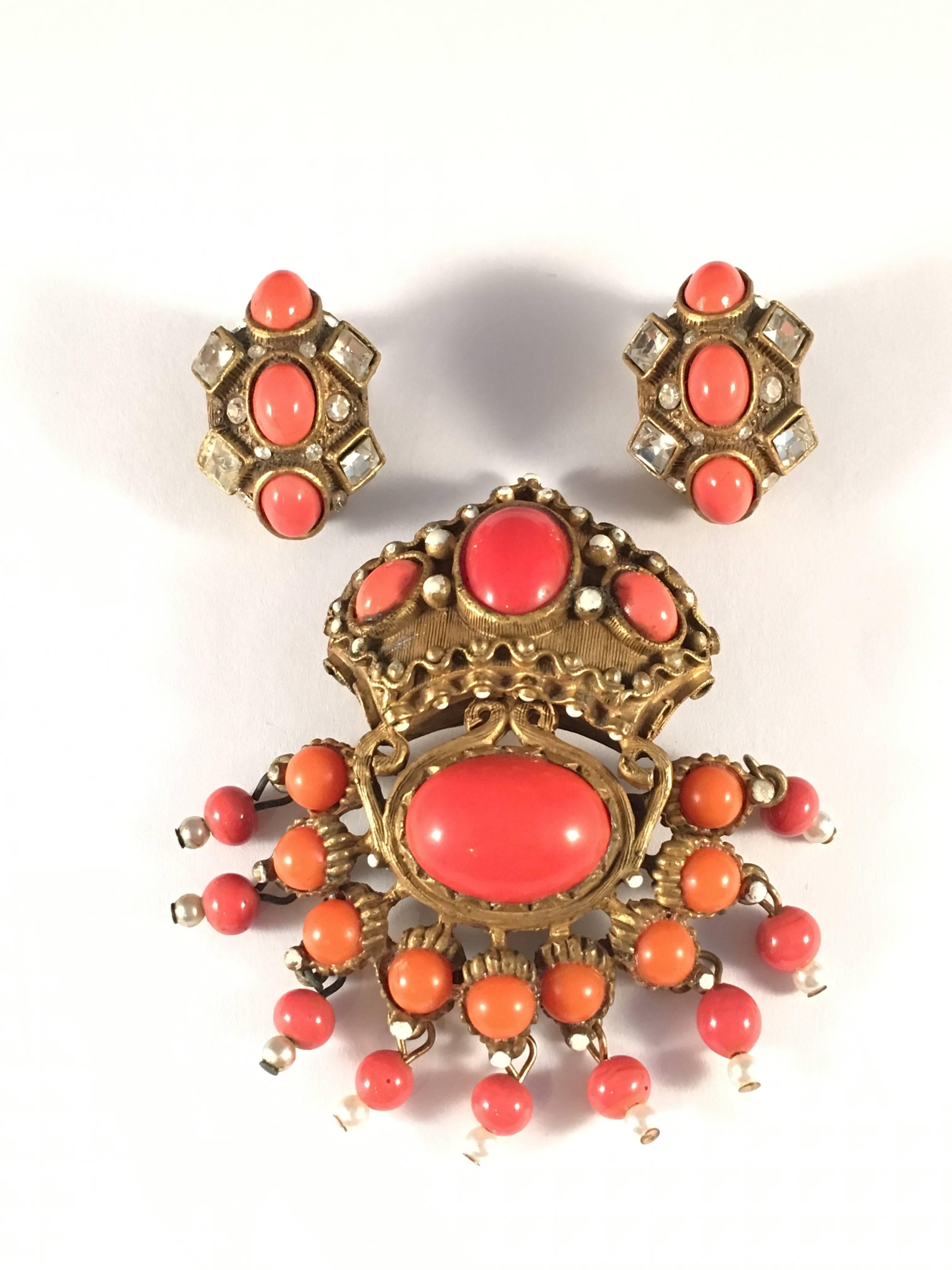 This is a rare and collectible 1960s Kenneth Jay Lane brooch and earrings set. It is designed with coral colored glass stones set in an antiqued gold-tone setting. The brooch has dangling coral beads with a smaller faux pearl dangling at the end of
