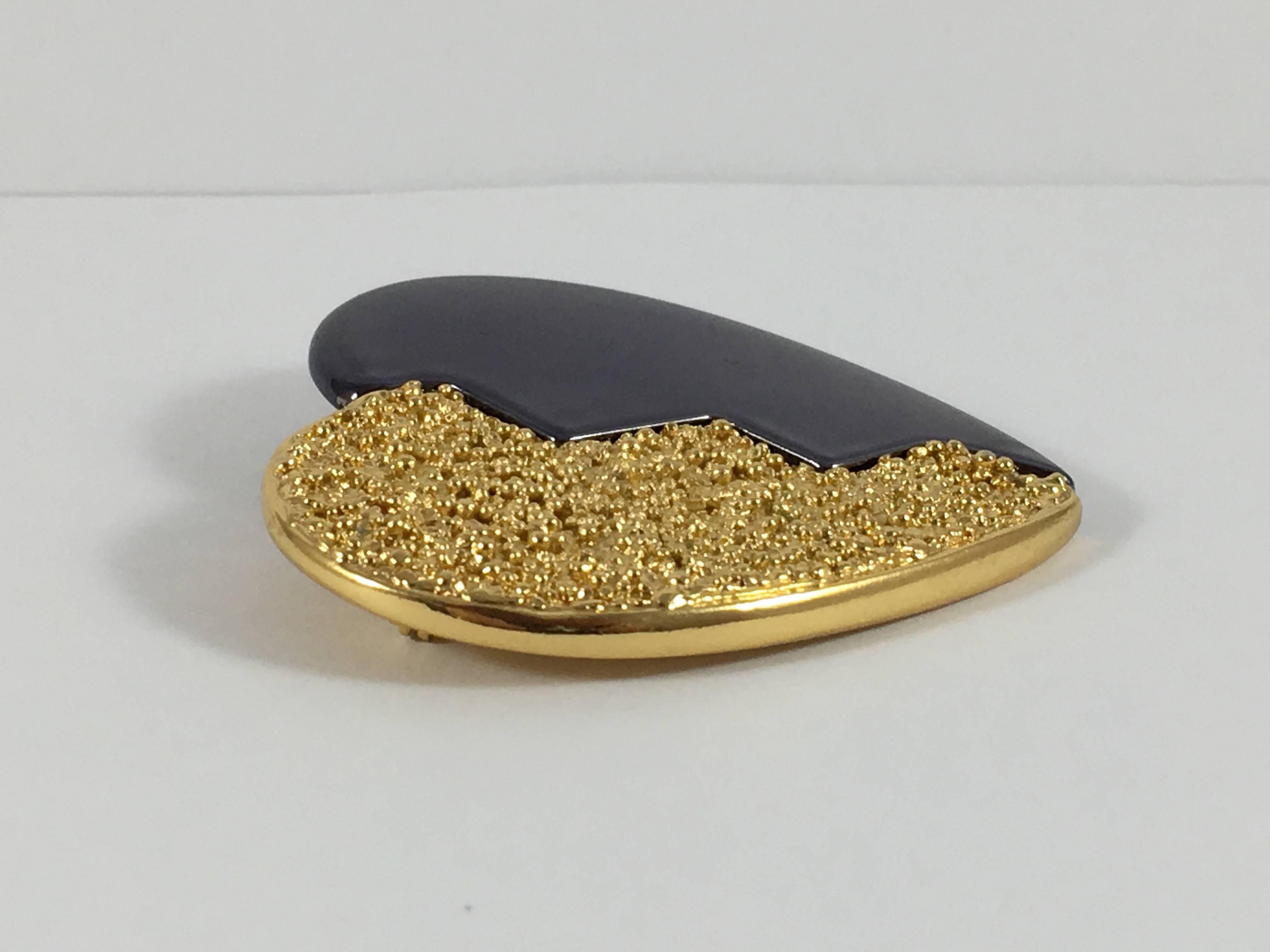 This is an Yves Saint Laurent heart brooch from the 1980s. It has a zig zag down the center. One half is a textured gold-tone metal and the other half is a smooth dark grey color.  It measures 2 1/16