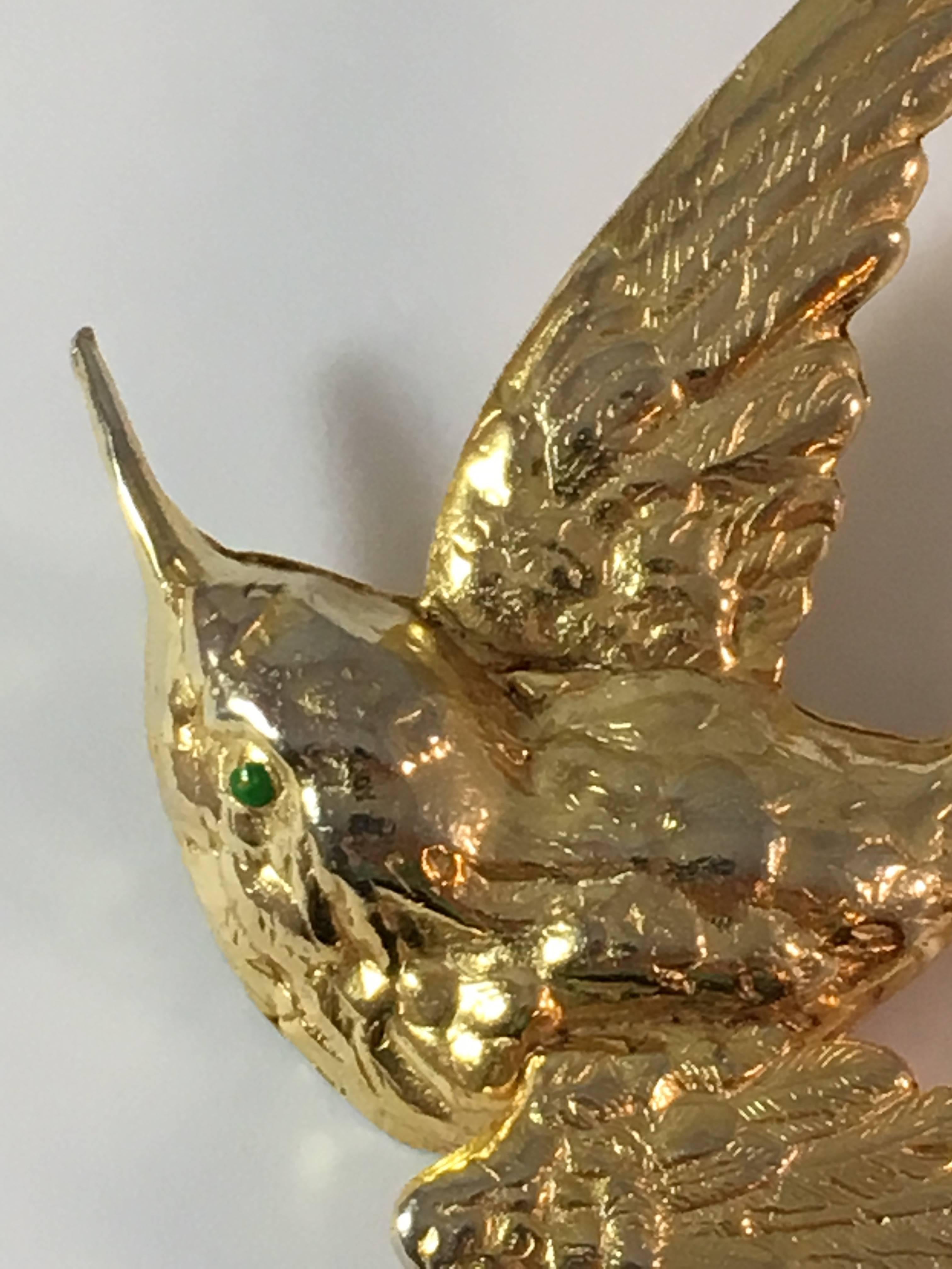 This is a beautifully made 1950s hummingbird brooch from Cadoro. It measures 2 3/4" wide by 2 5/8" tall. It is made from a gold-tone metal and has a green enamel eye. It is marked 'Cadoro' on the back in an oval plaque. It is in excellent