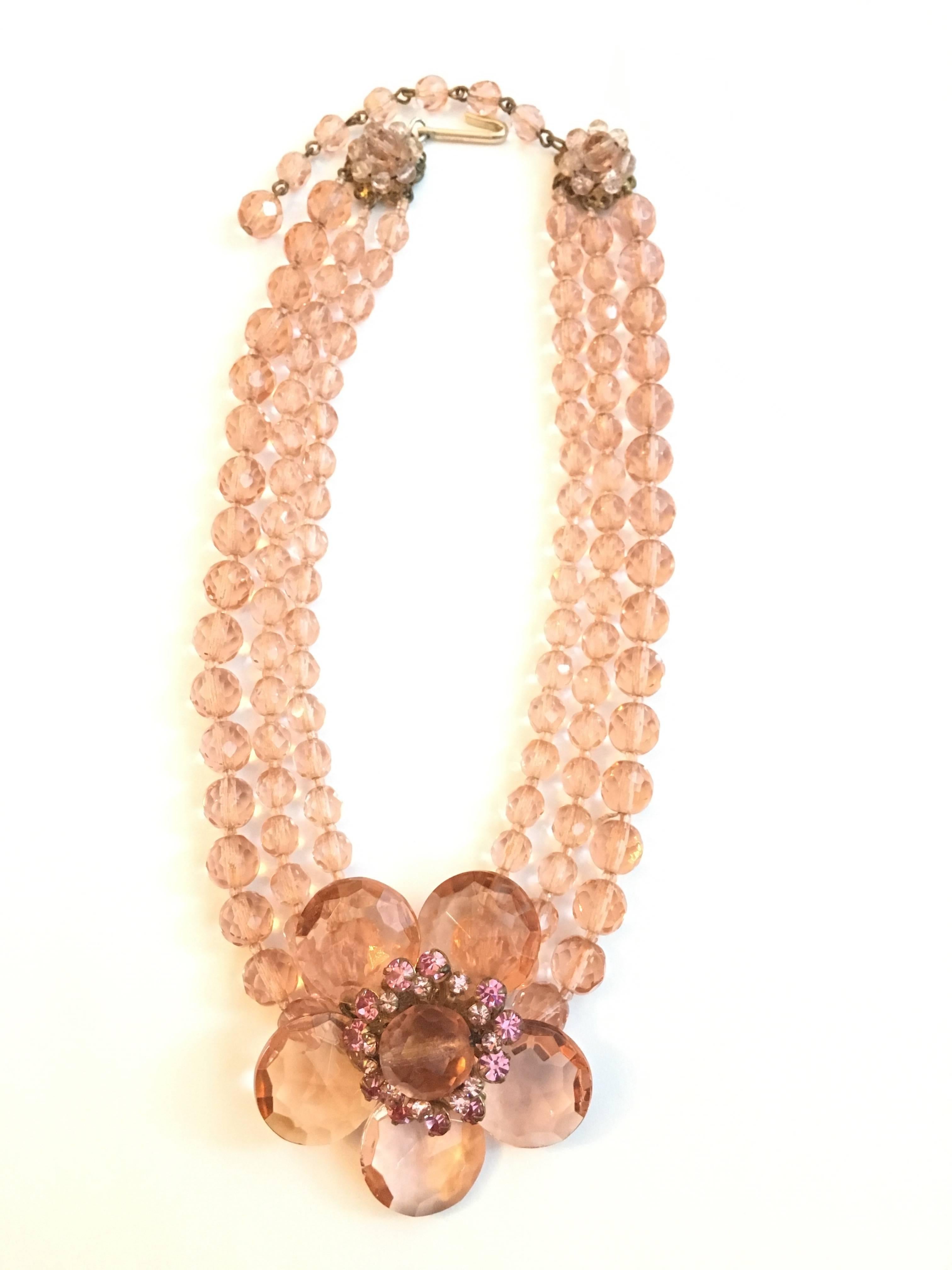 This beautiful three strand Miriam Haskell choker necklace is made up of pale peachy pink bohemian glass crystals. It features a large five petaled flower in the center which is mounted on a classic Haskell gold plated filigree disk. The flower