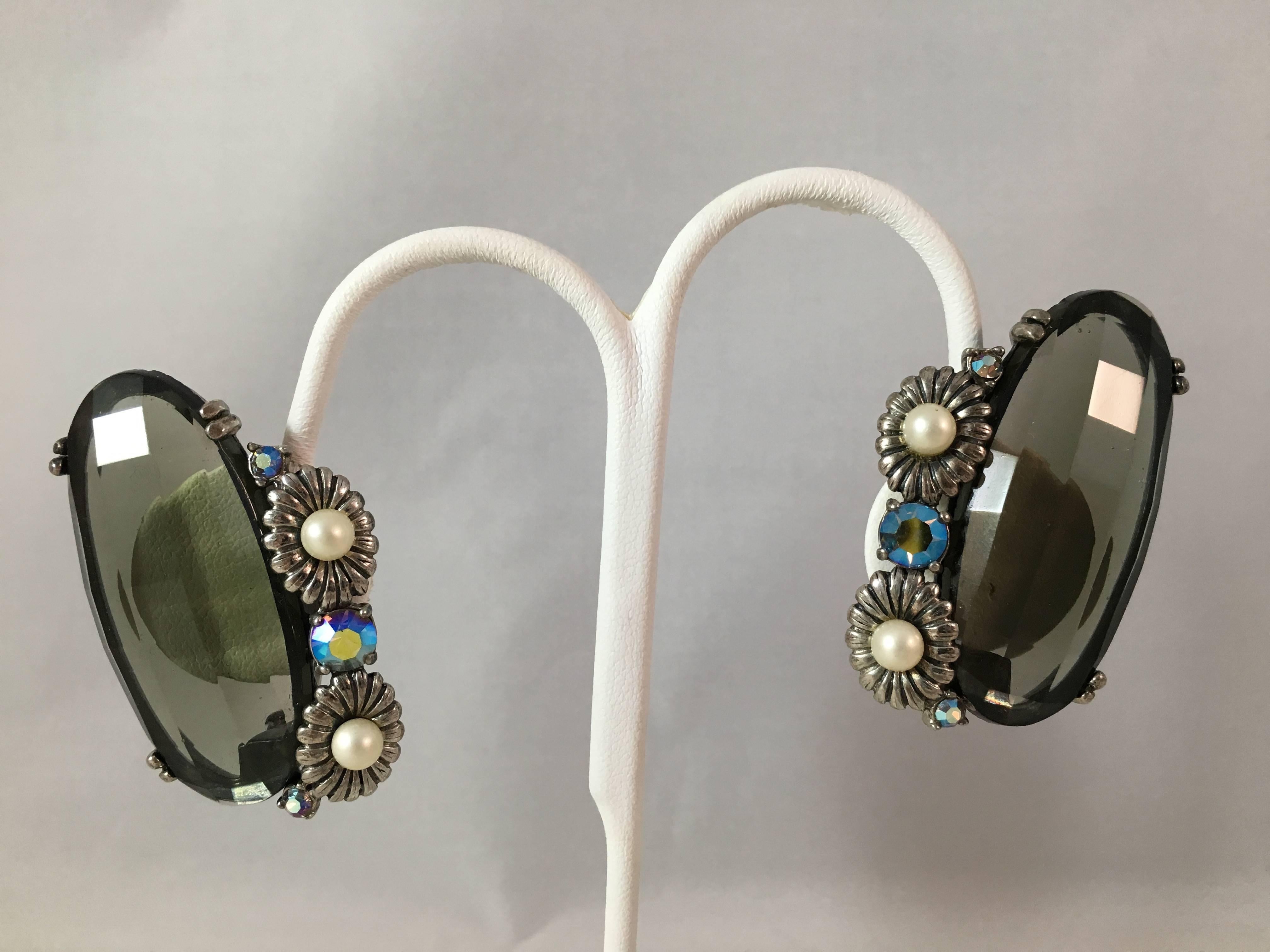These are huge Schiaparelli clip-on earrings from the 1940s. Each earring is  made up of a large faceted grey topaz glass stone set with silver-tone metal flowers with faux pearl centers and aurora borealis stones. The earrings are set in a