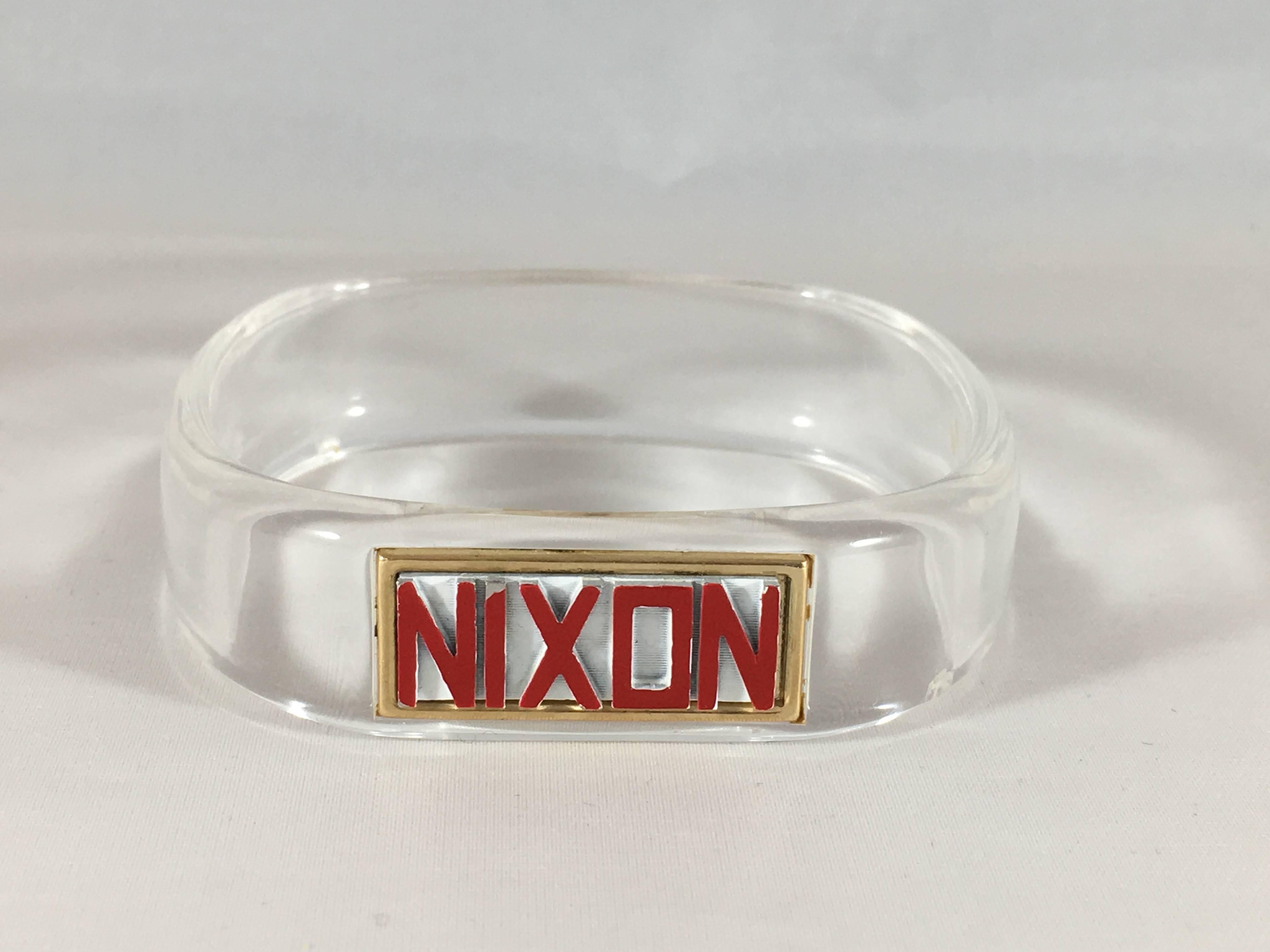 This is a collectible 1960s Trifari clear lucite 'Nixon' bangle bracelet. It has a red and white enameled gold-tone plate inserted into it that reads, 'Nixon'. It was most likely made in 1968 for the Presidential campaign. It has a great mid-century