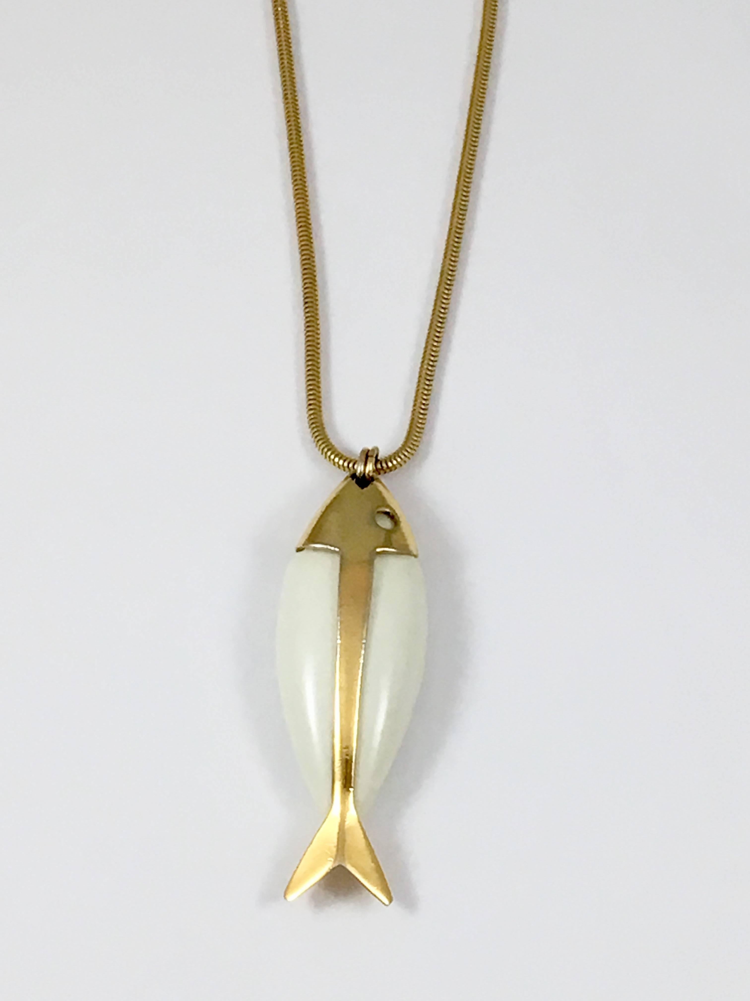 This Trifari fish pendant necklace is from the 1970s. It features a white resin and gold toned metal fish pendant measuring 3 1/2 inches long x 1 1/8 inches wide. It hangs from a gold toned snake chain measuring 34 inches long. The necklace is in