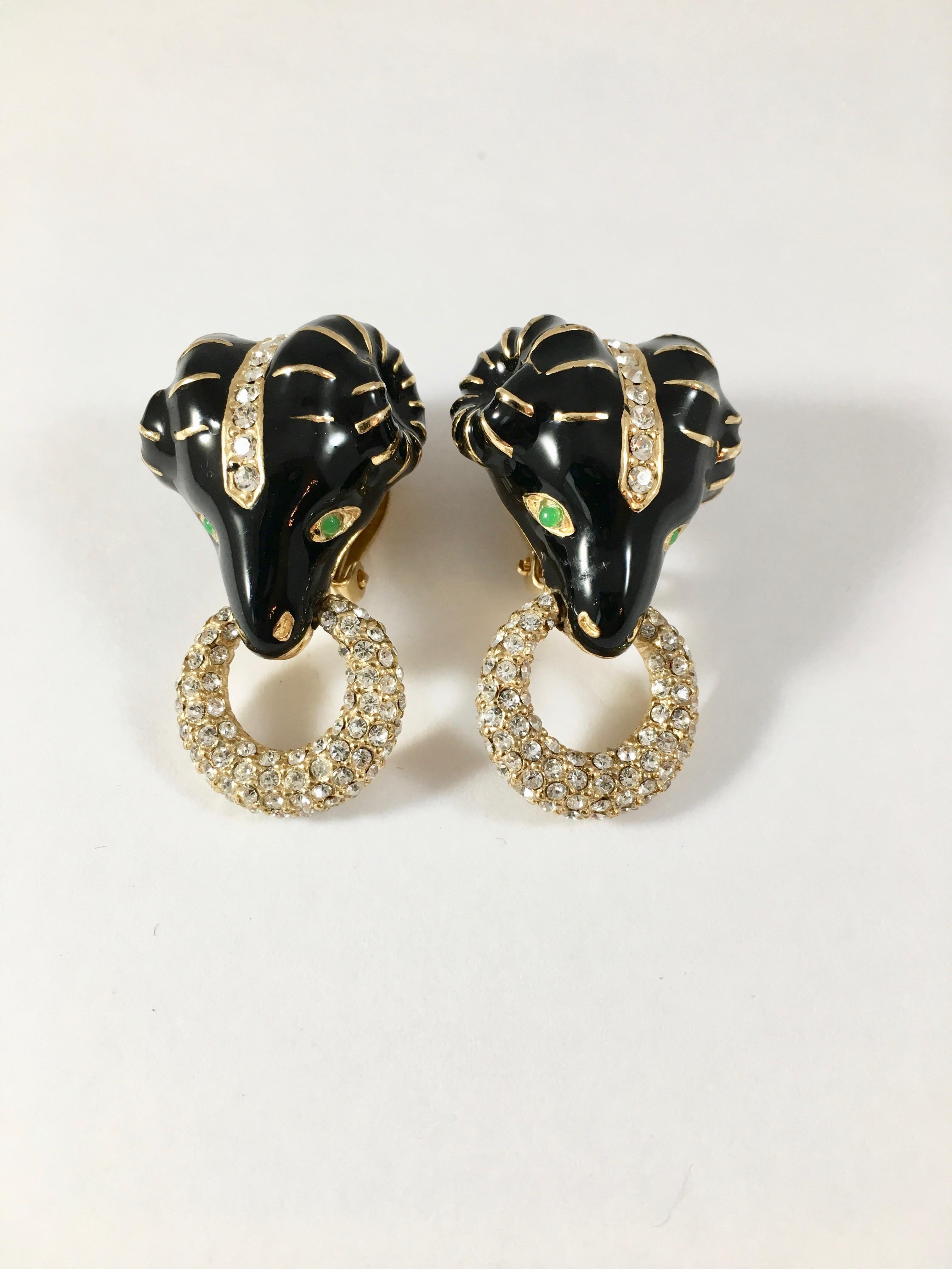 This is a pair of 1980s Ciner rams head clip-on earrings. They are covered in black enamel and have green glass eyes and clear rhinestones imbedded in their heads. Each ram has a ring covered in clear rhinestones in their mouths. They are marked