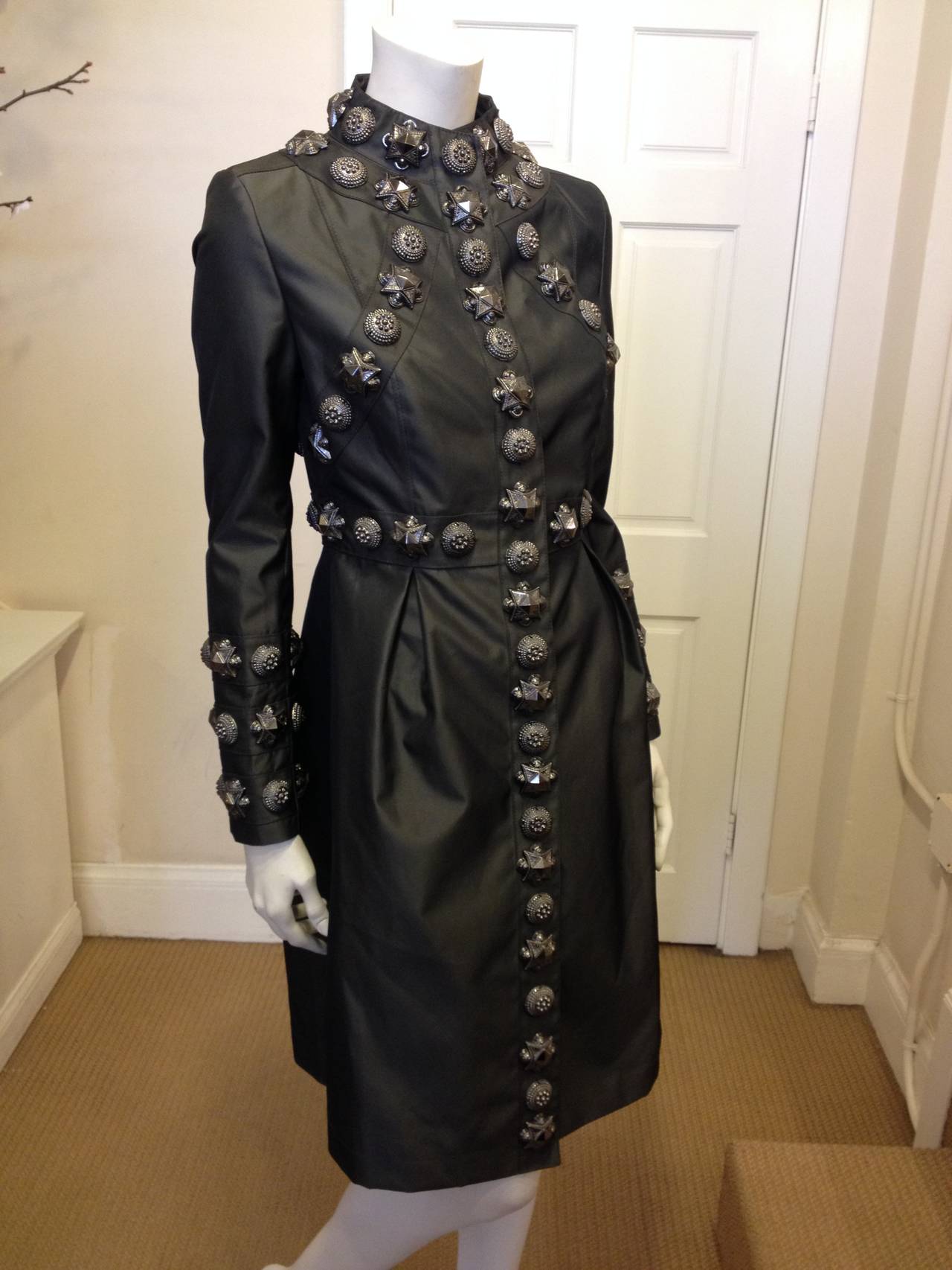 This coat is over the top! In glossy gunmetal grey taffeta, it features big and intricately embossed pewter-colored studs marching across the chest like a British coat of arms. The high neck, long sleeves, and mid-calf hemline add an angularity and