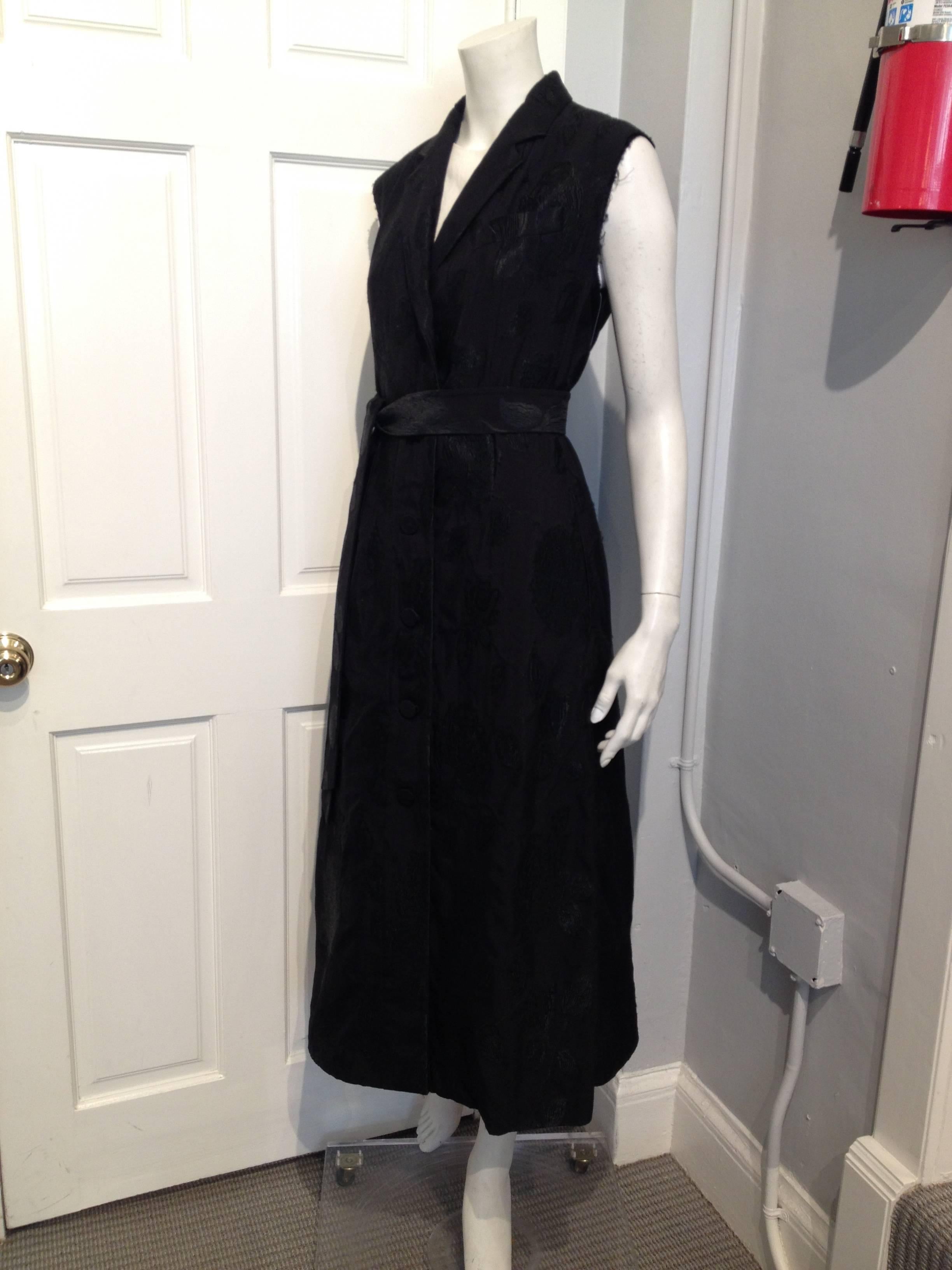 Gorgeous black-on-black floral brocade material cut into a long sleeveless dress coat with lapel collars, brocade-covered buttons running down the front, frayed sleeves, and a below-the-knee hemline. Belt is removable.