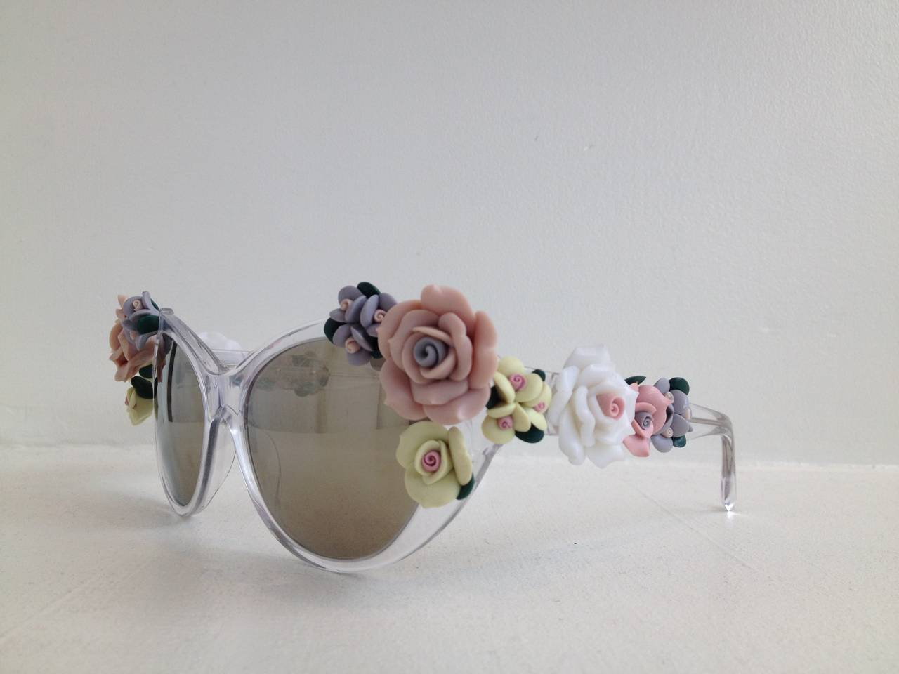 Absolutely fabulous! These glasses do a lot more than keep the sun out. Crystal clear frames, in a retro cat eye shape, are decorated on either temple with bouquets of delicate, sculptural, pastel rosettes. The lenses have a deep honey-colored