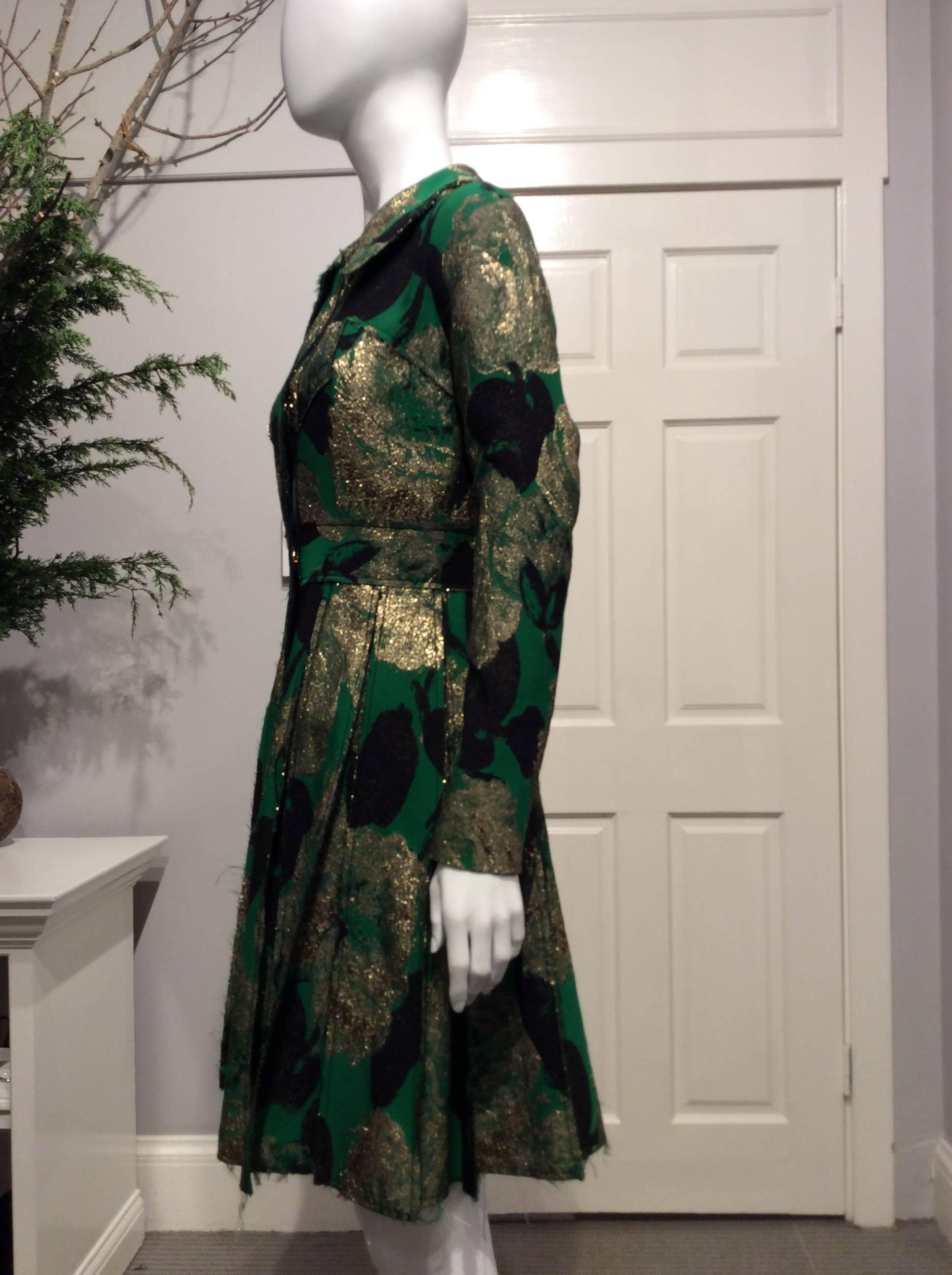 Coat-dress in a vibrant green, gold and black abstract pattern with sewn flat pleats. It closes with six shiny oversized snaps. The waist is defined by a 2.25in wide band in the same fabric and reinforced on the inside with grosgrain. It has two