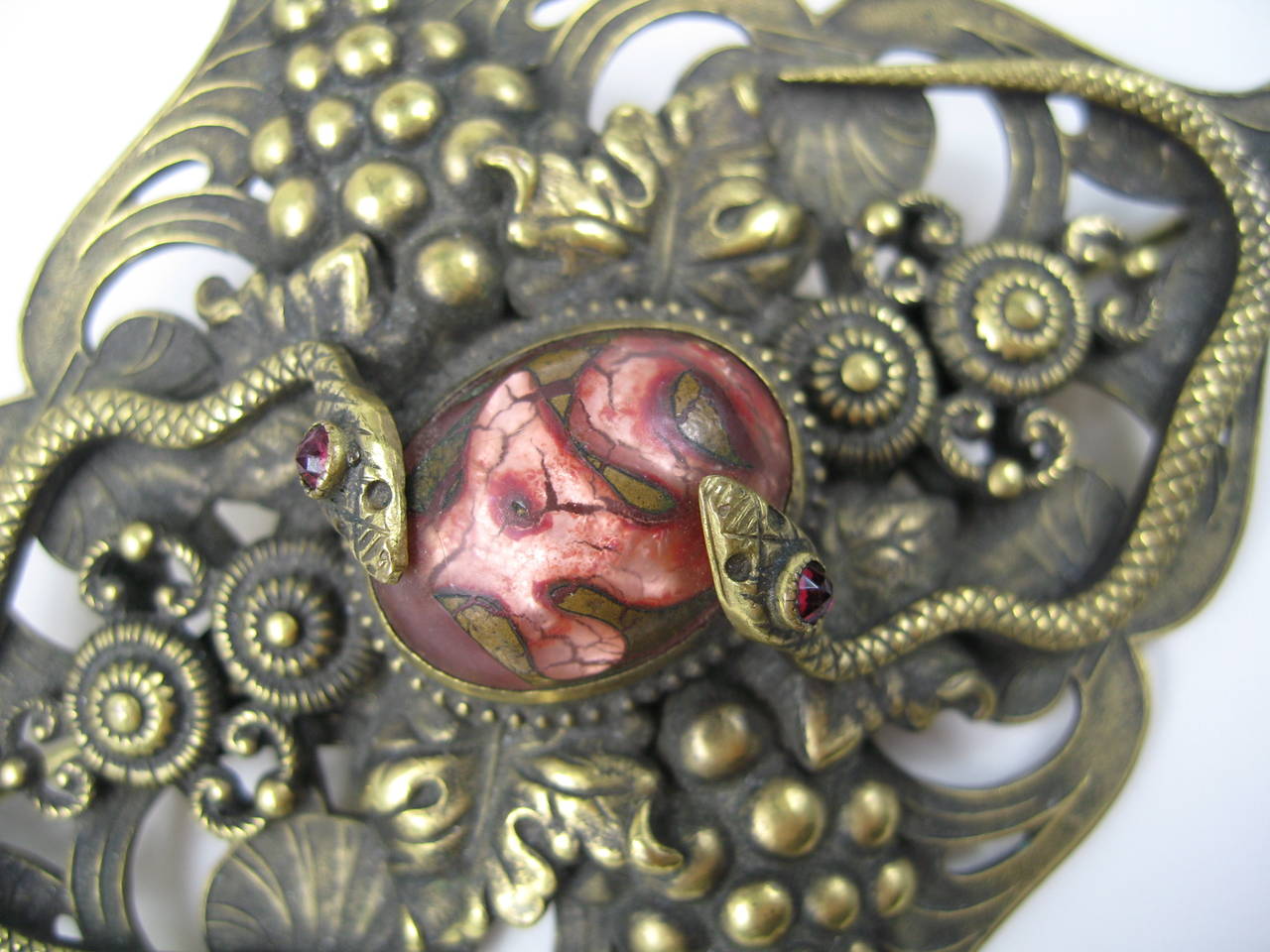 Wonderful Victorian brooch with double headed snake and bezel set center stone. The Pink Cabochon has gold glass blown into it to create a vine or snake like effect. Two snakes circle this stone. Early C back pin back. Made of gilt heavy brass.