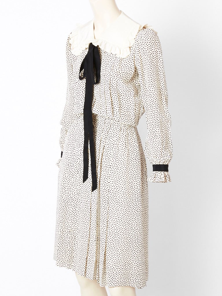 Yves Saint Laurent, ivory silk day dress with black tiny polka dots.
Dress has a large square ivory collar, edged in ruffles. Buttons go down the middle front and end at the lower hip. Bodice is pleated in the center front, with the same pleats