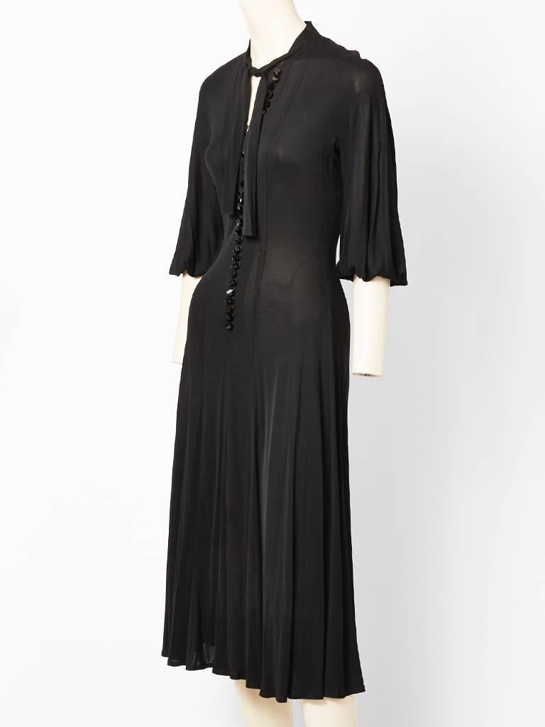 Jean Muir, matte jersey, 70's day dress having a fitted bodice, with a paneled skirt that flares out. Dress is fastened up the center front with tiny, small, round black buttons that are secured with loops. Neckline is collarless with a tie, that