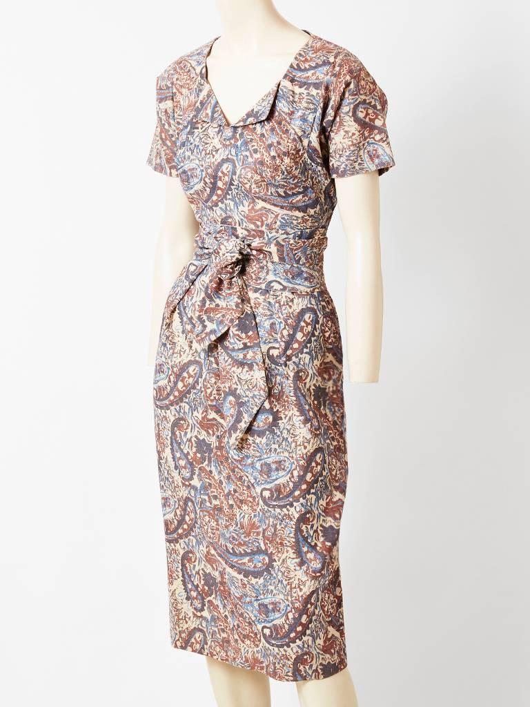 Claire McCardell, muted tones,  fitted day dress having a paisley-like pattern.
Dress is cinched at the waist with an obi-like sash. Short sleeves with a scoop neckline have a shallow center slit.