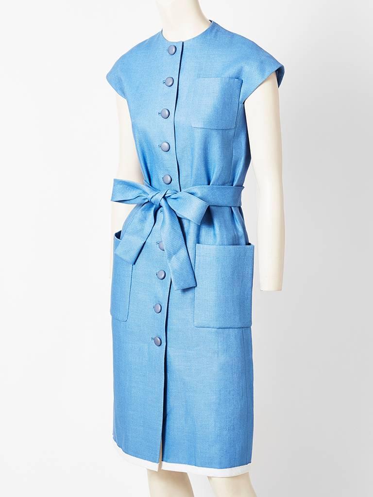 Norman Norell, cornflower blue, moygashel linen, capped sleeves, day dress, having a sheath silhouette, that buttons in the front with a self belt. Single Breast and hip pockets are the only embellishment on the dress. Dress is old stock with a dust