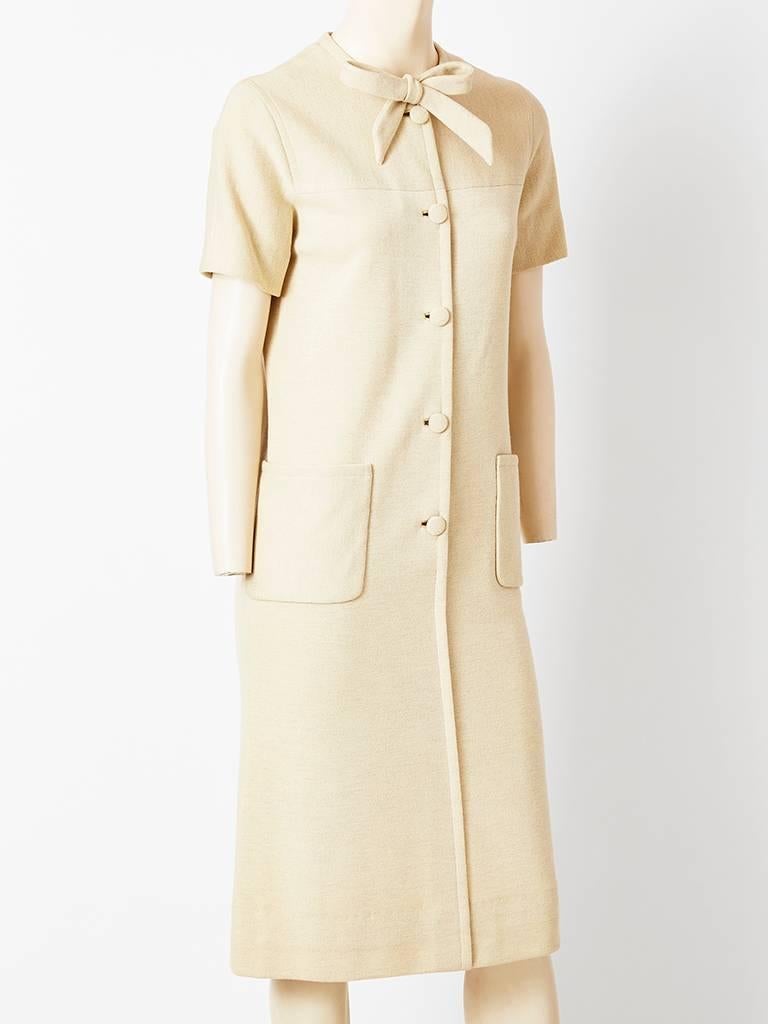 Norman Norell, oatmeal tone, wool knit, sheath, having buttons closing down the middle front, short sleeves, a rolled neck with a front bow and visible deep patch pockets.