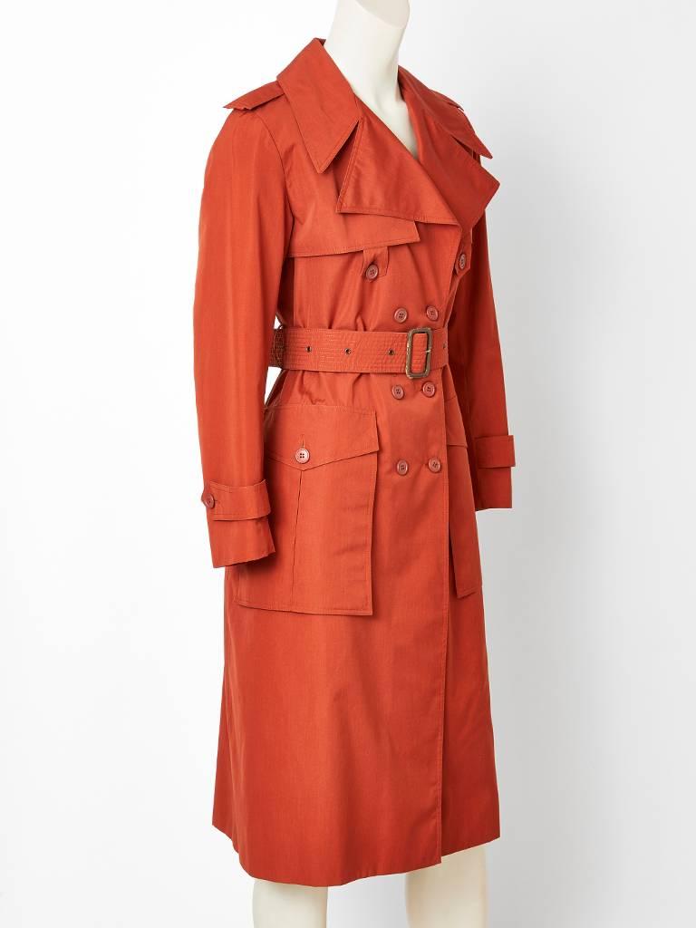 Yves Saint Laurent, Rive Gauche,  fitted, rust tone,  double breasted, cotton poplin,  trench, having wide lapels, a stitched belt,  epaulettes and deep pockets with buttoned flaps. 