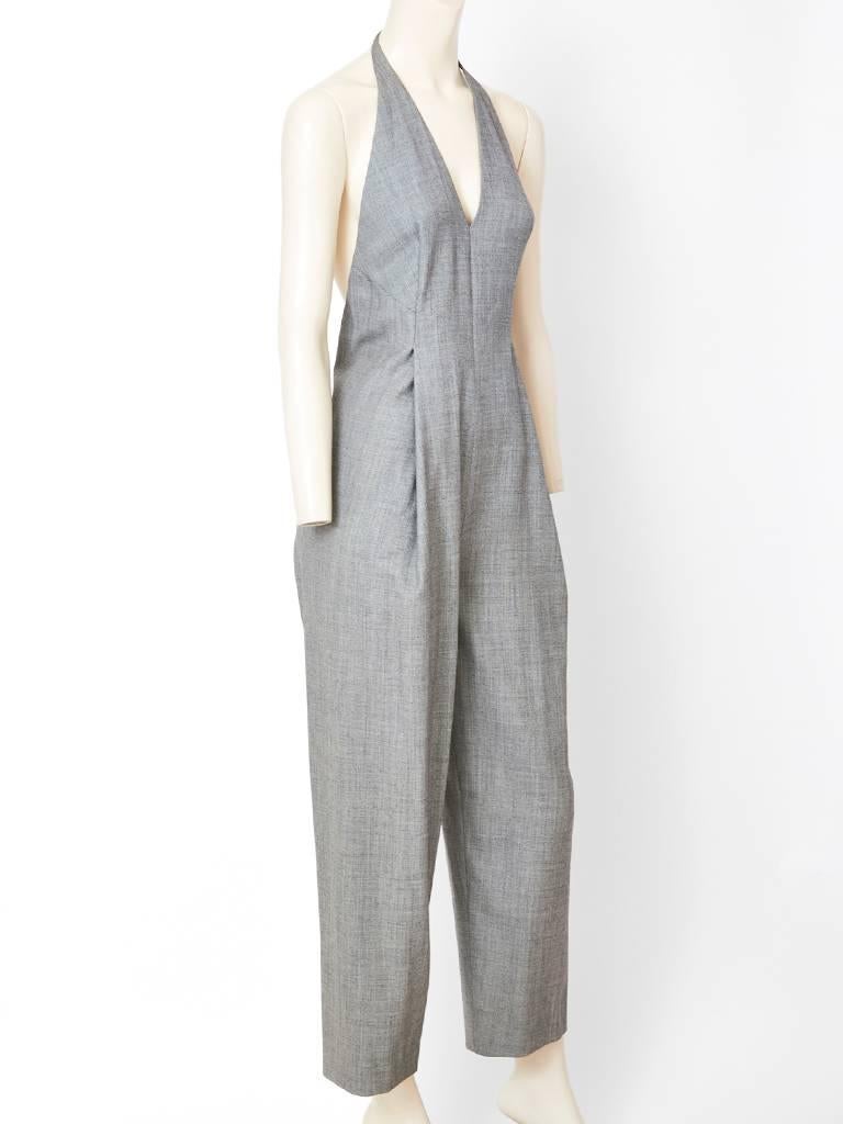 Geoffrey Beene, fine wool jumpsuit in a gray menswear fabrication, having a halter neckline, and open back. The front has a deep V. There is an accompanying cropped  jacket with a cap sleeve and a small pointed collar. 