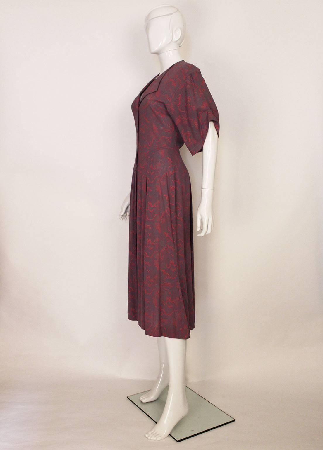 An elegant and easy to wear dress by British designer Jean Muir.
The dress is made of cotton with a wonderful pink and purple print, and the buttons are hand painted.The dress is fitted to the body, and the skirt flares at the hip. There is a full