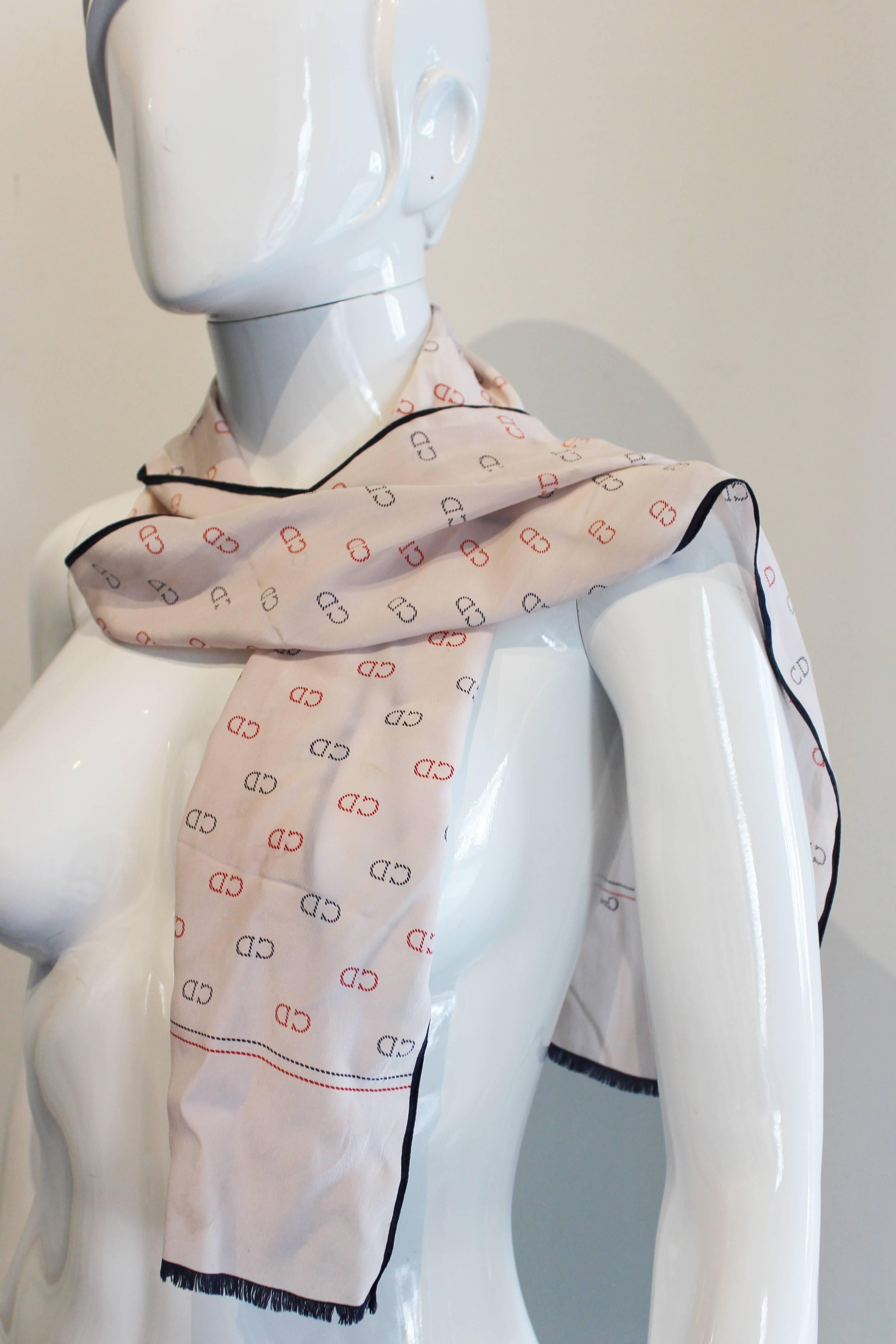 A chic silk scarf by Christian Dior.This silk scarf  has a very pale grey /pink background  with a black vertical border and fringing at either end.The body of the scarf has the CD logo in red and black printed throughout,and here is a red line and