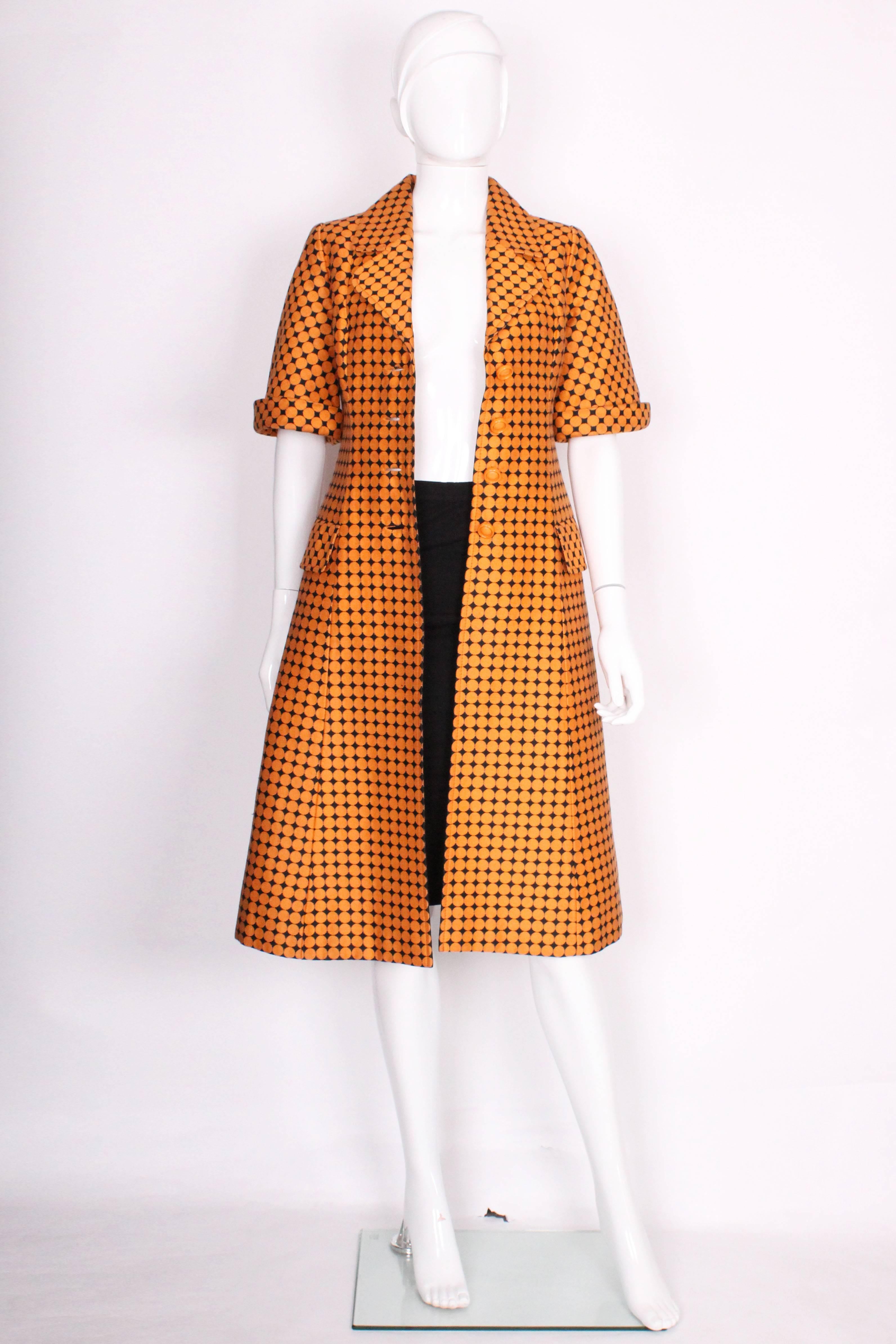 A chic and bold coat dress made for Marshall and Snelgrove by IFL Couture in Switzerland. The coat has a black background and large orange dots that. It has a large double lapel collar and 4 round orange buttons down the center that closes the coat