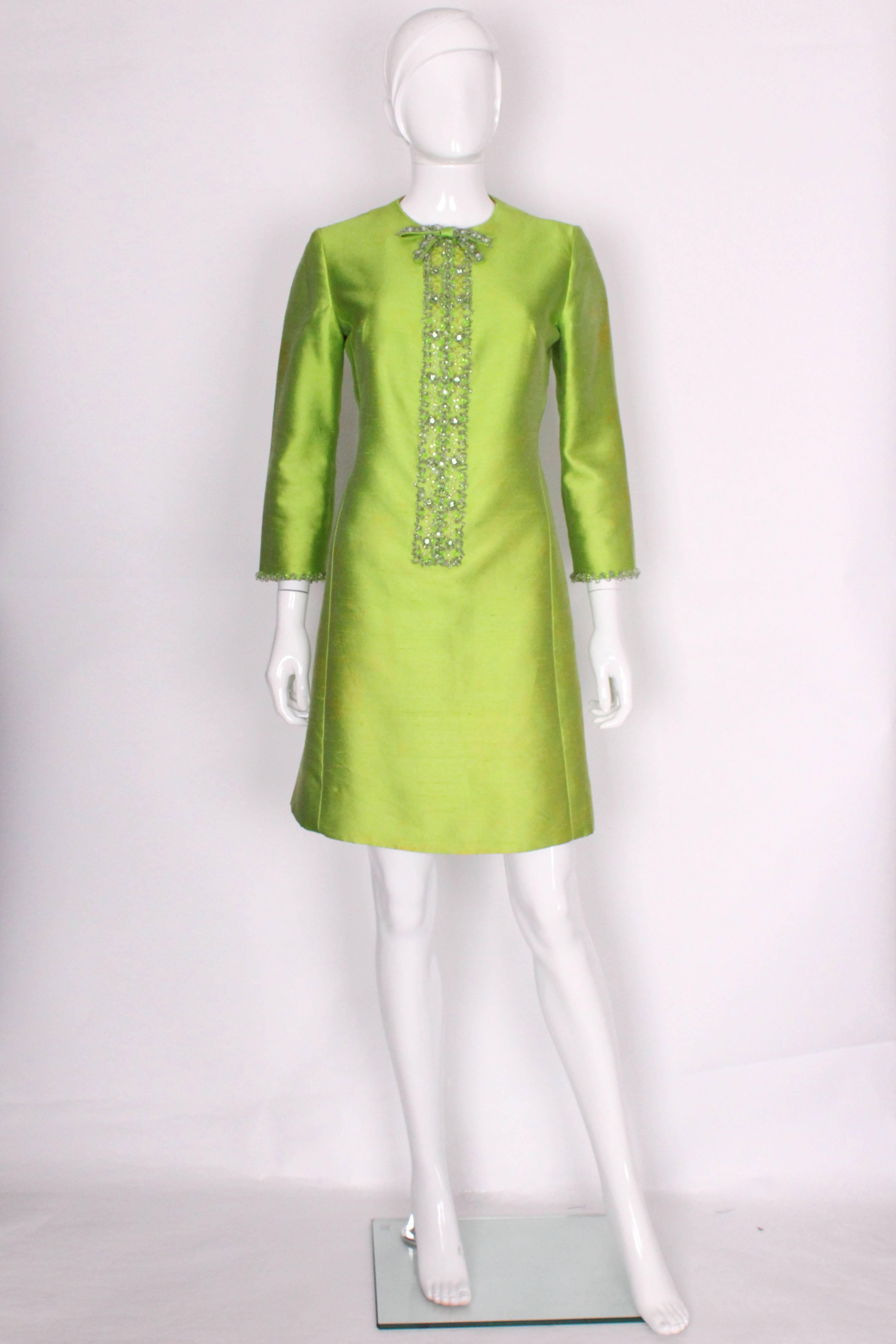 A great bold cocktail dress from the 1960s. In a stunning, eye-catching bright green slub silk with wonderful beadwork to the front and cuffs. The bib detail has a lovely bow at the top that is also embellished in beadwork. The colouring of the