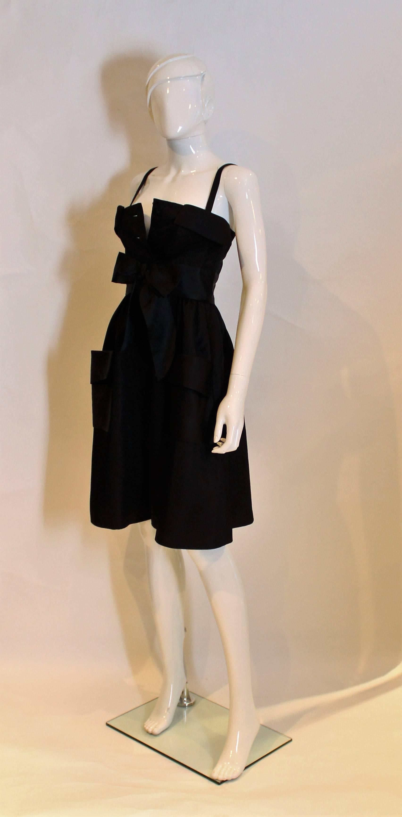 A delightful  little black dress by Geoffrey Beene. The dress has a fold over top on the bodice, 8 button front opening, bow at the waist and two pockets on the skirt front.