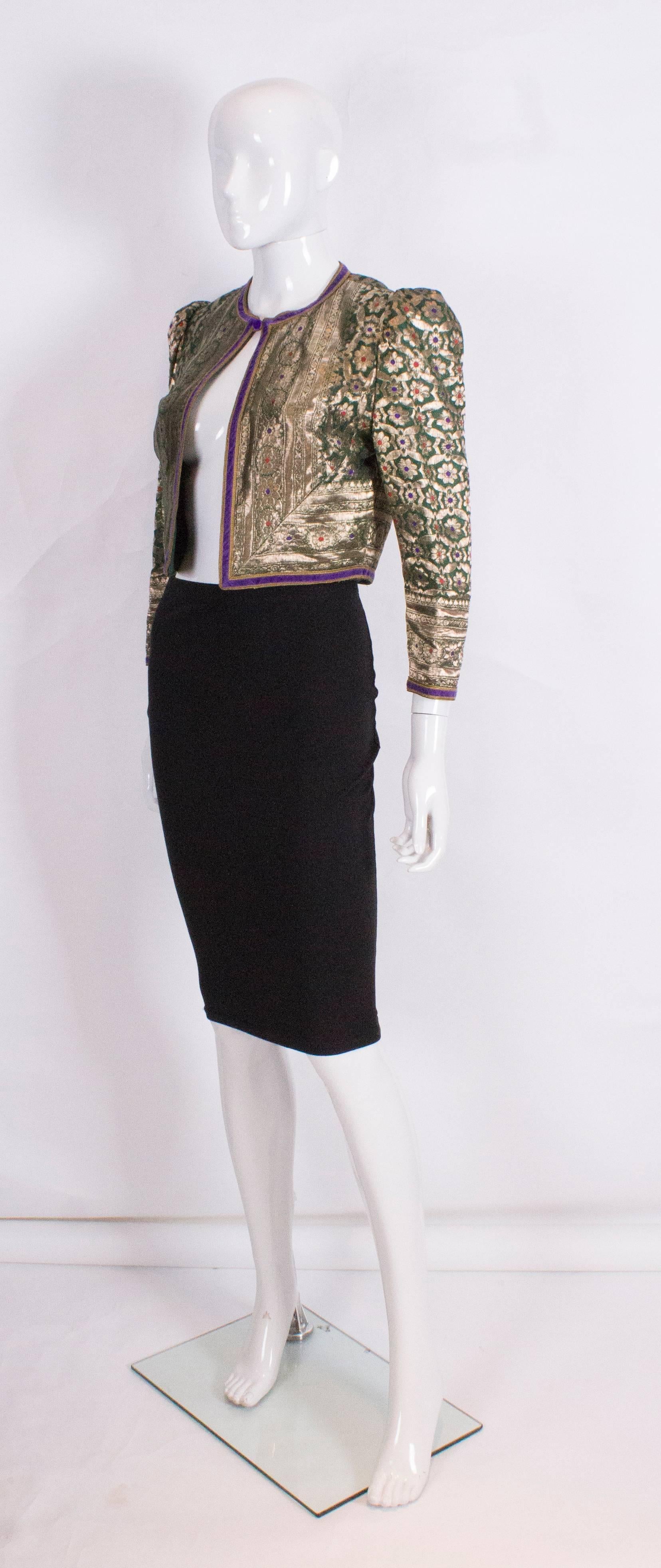 A great vintage jacket  by Regamus. The jacket has a green background with gold thread and a floral design with a purple velvet border. The jacket is collarless, with a one button fastening, gathered shoulders and is fully lined.