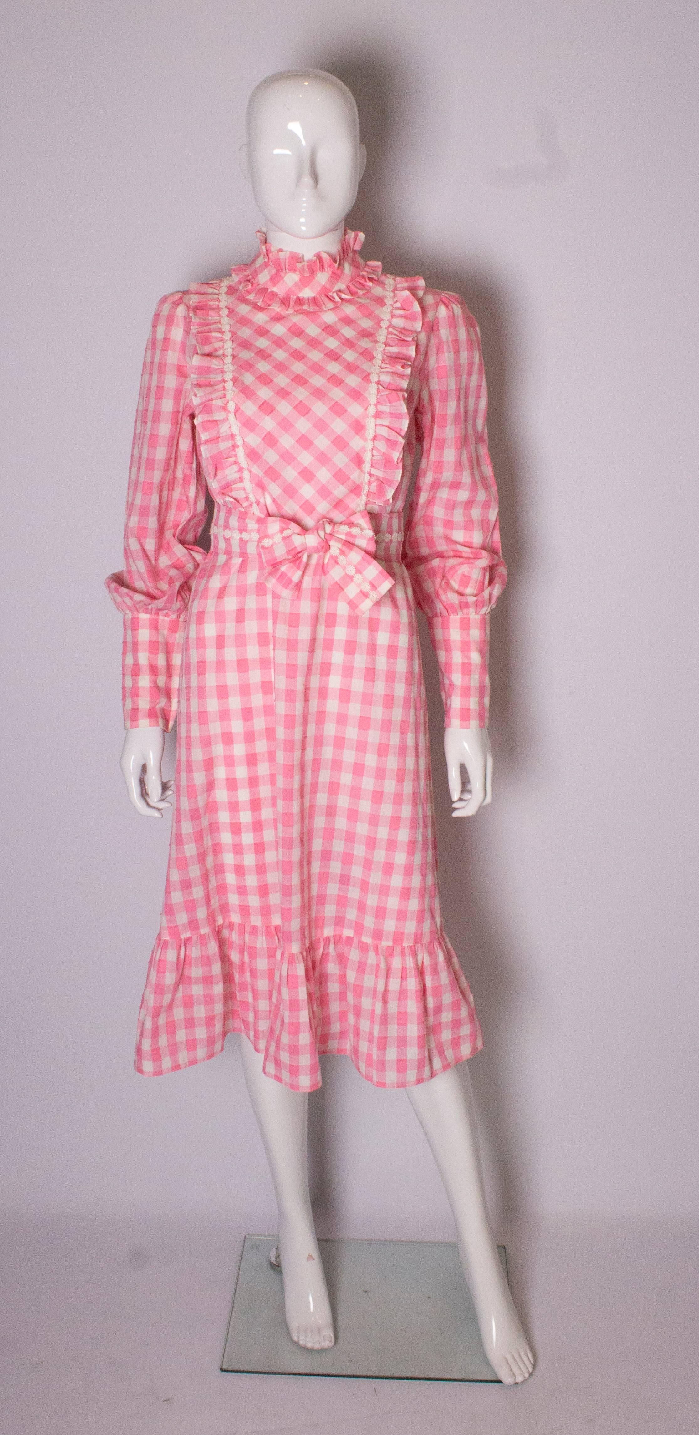 A fun dress for Spring, by British designer Susan Small.. The dress is made of a pink and white check fabric, with some texture detail on the pink squares.It has a stand up collar with two rows of frills, and a yoke with white flower detail and