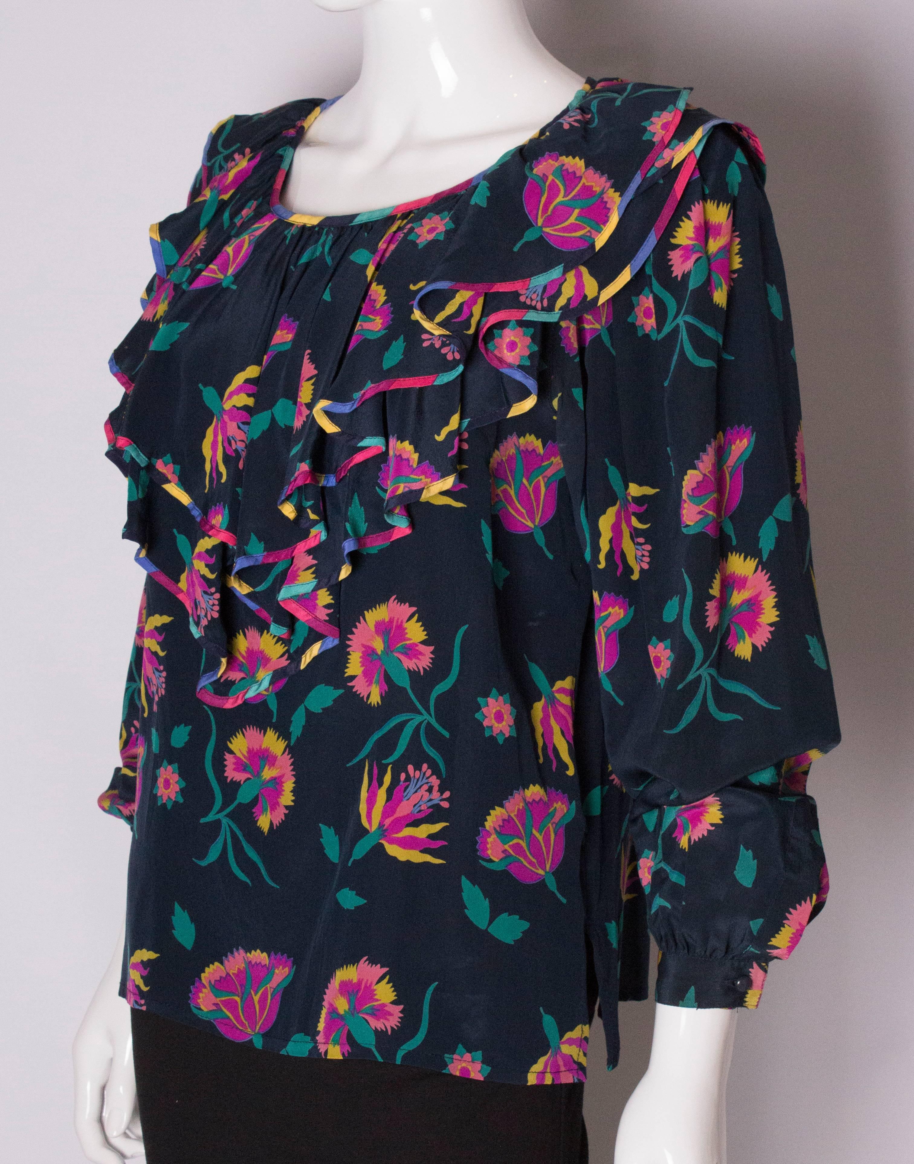 A pretty silk top by Escada. The top has  a blue background, with a  pink , maroon and green design. It has a v neckline with frill around the neckline, and single button cuffs. It has a 3 1/2 '' slit at either side. 