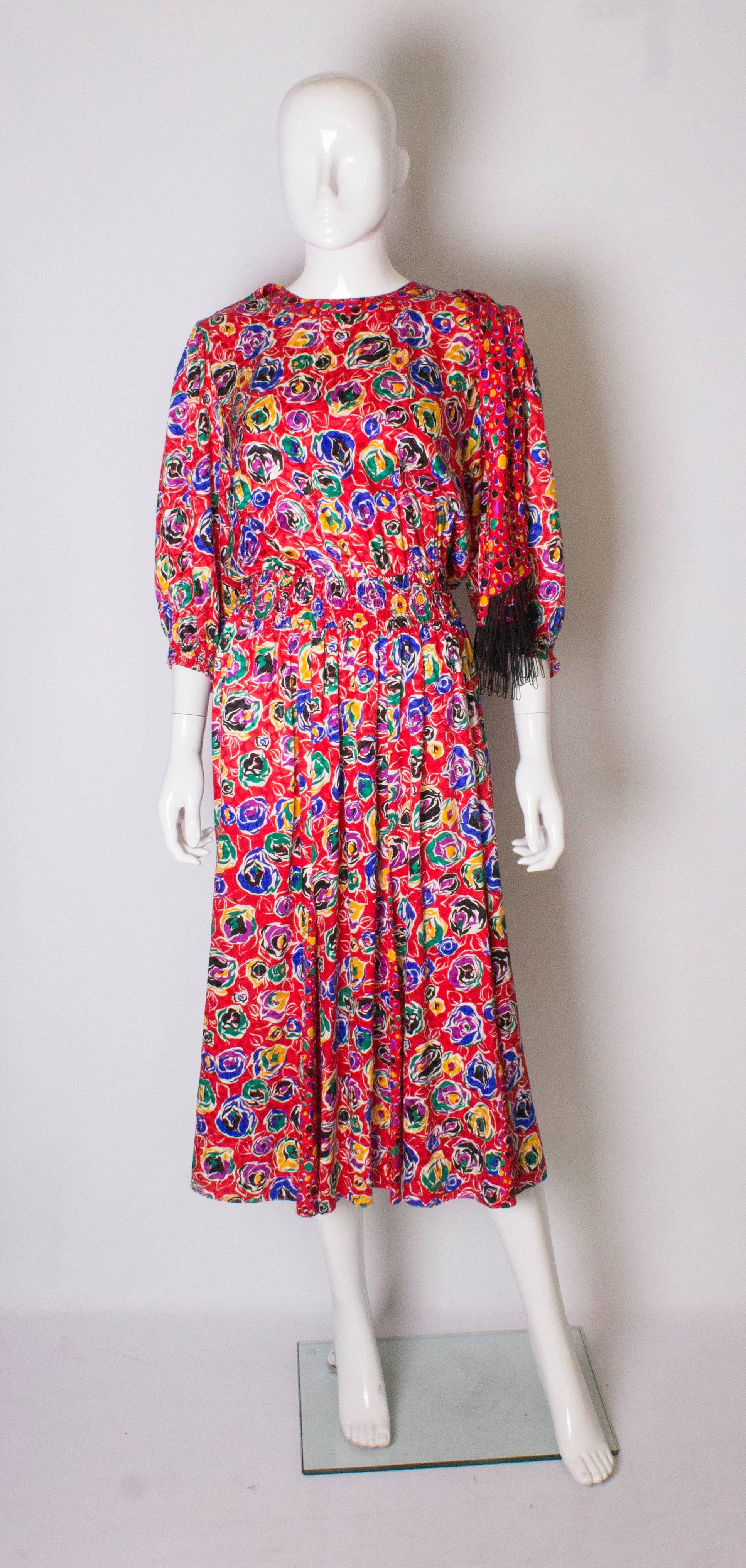 An easy to wear dress by Kanga /Dale Tyron. The dress has a red background with a multi-coloured abstract floral print. It has a round neckline. elbow length sleeves and an elasticated waist.   There are panels at the lower skirt, so the dress