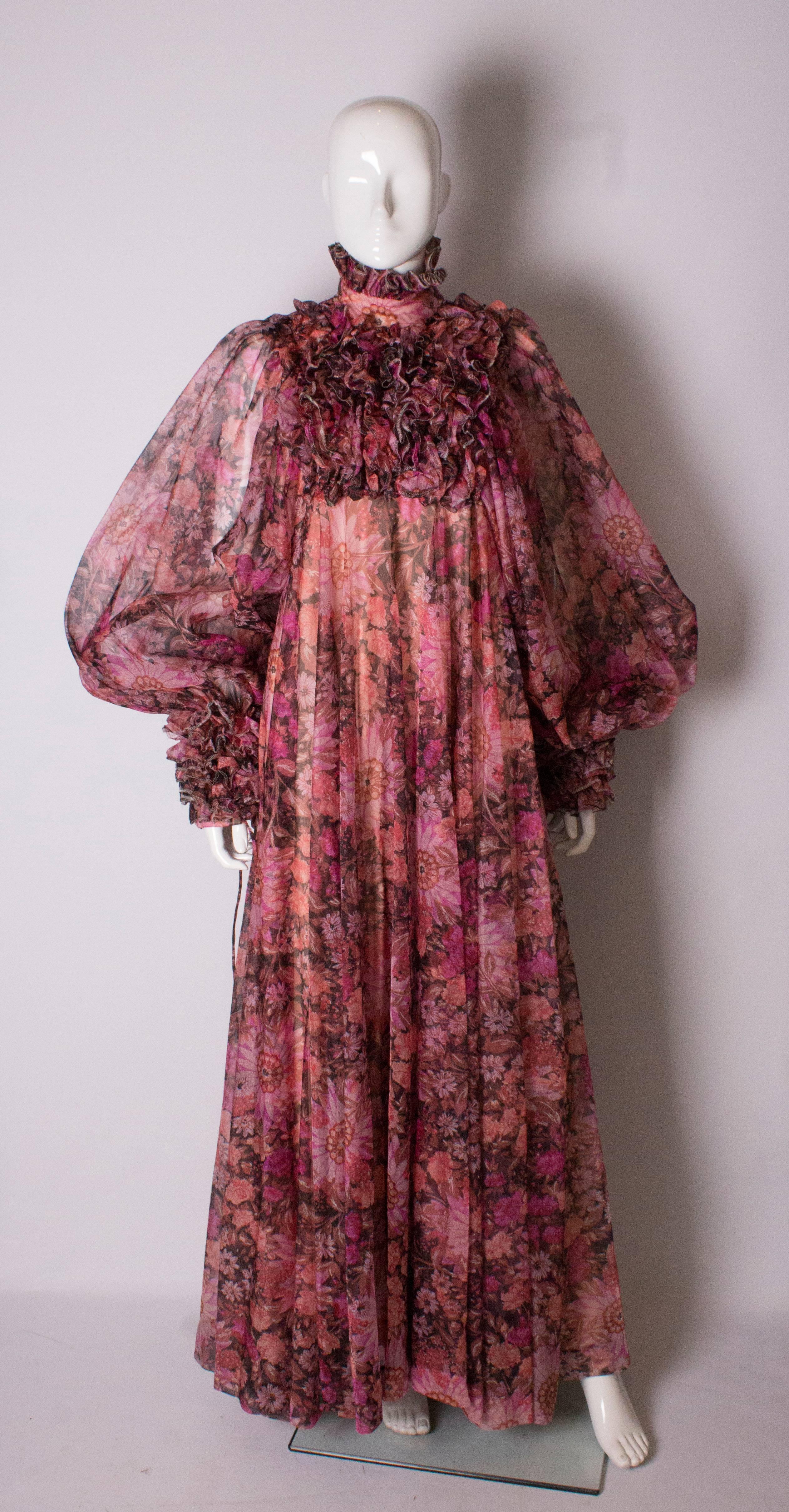 A stunning gown by Jean Varon. The dress is a bright floral print of shades of pink and purple on a black background. The dress has a stand up collar with frill trim ,and frill on the front., and the cuffs are wide with tie detail. The dress is