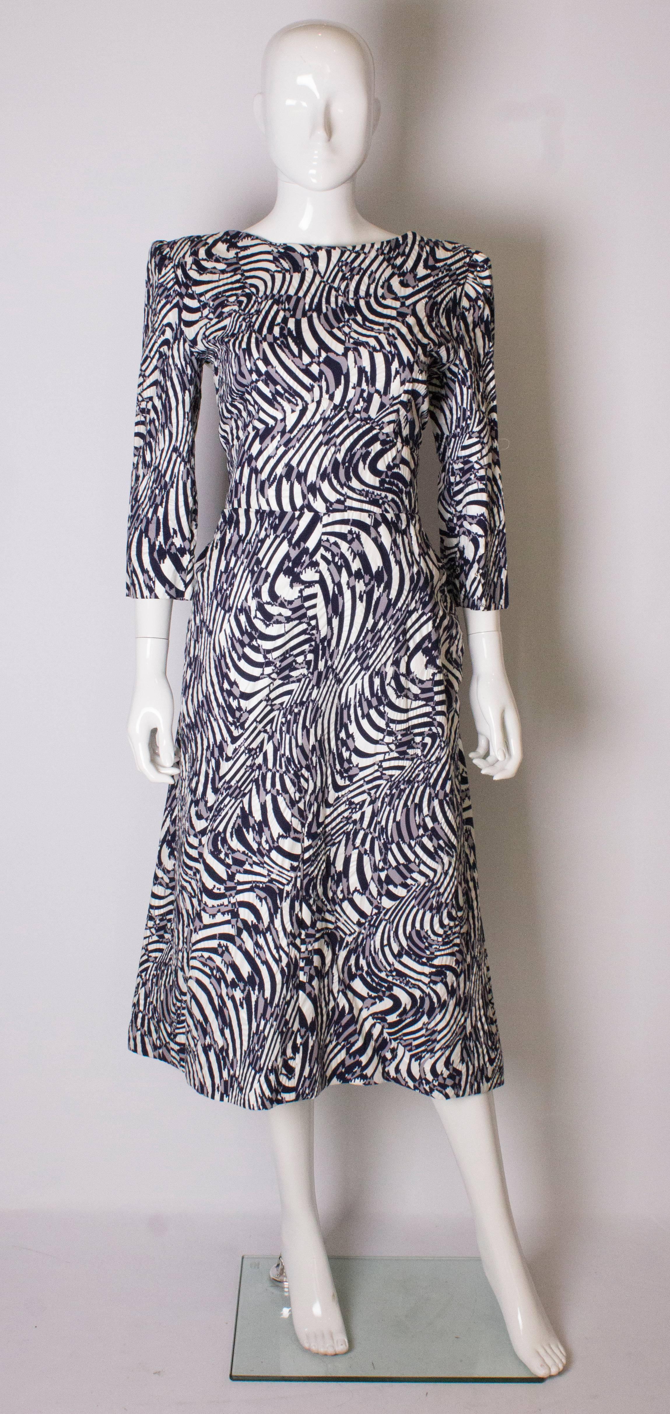 A great dress by Donald Campbell. The dress has a white background with a grey and blue print. It has a round neckline, elbow length sleeves, and a scoop neckline at the back. There is a  central back zip, a back bow and peplum. The sleeves narrow