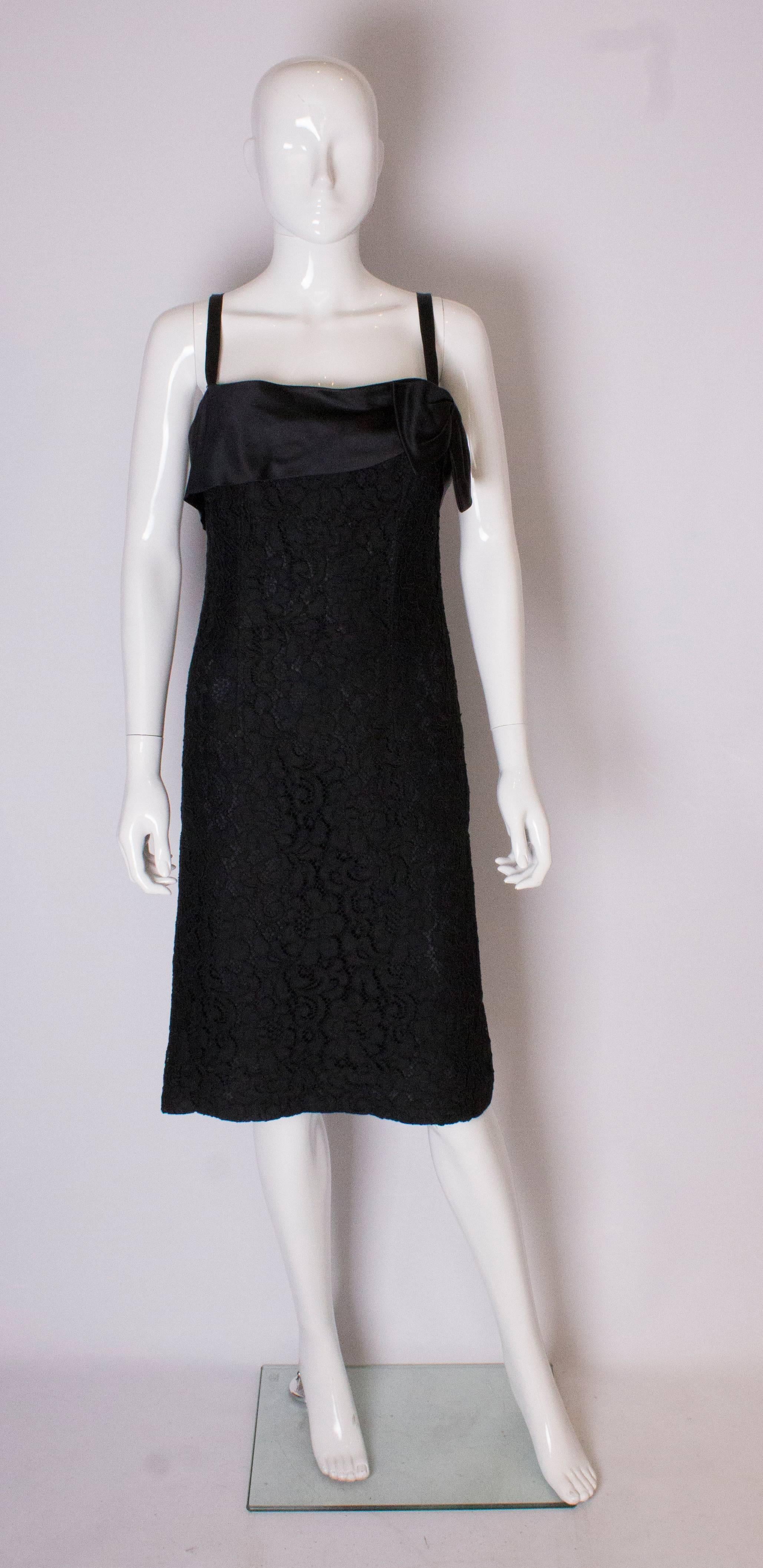 A chic  black lace dress. The body of the dress is black lace with a black lining underneath. There is a satin band and bow at bust leval ,ribbon straps and a central back zip.