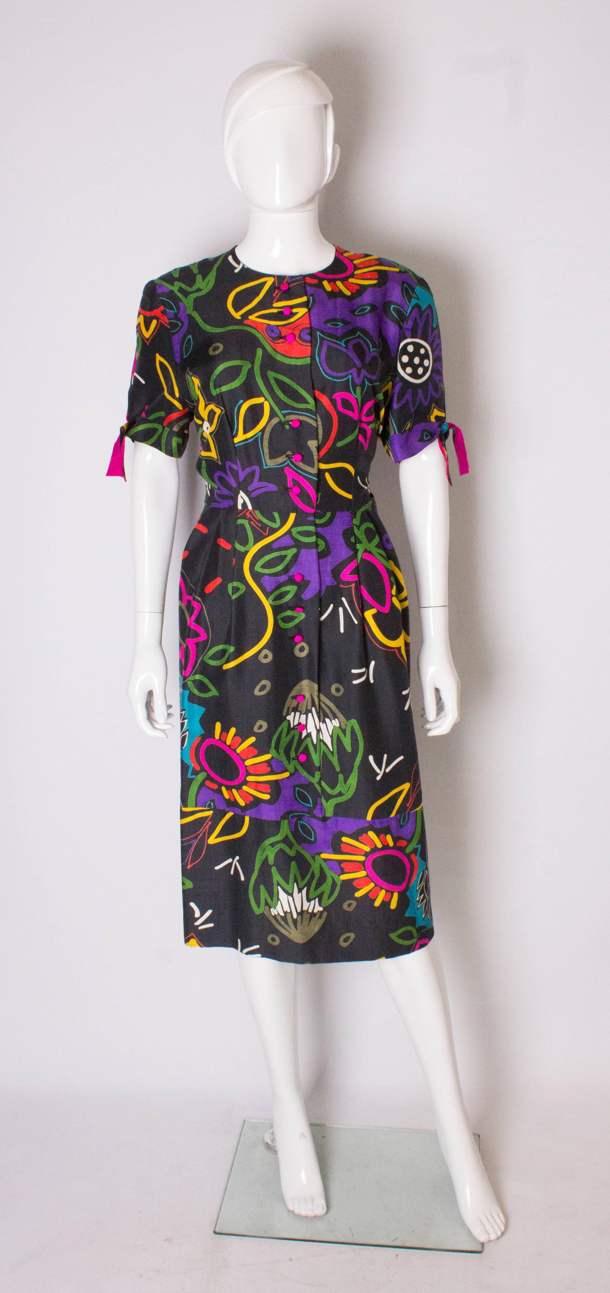 A chic day dress by British designer Donald Campbell. The silk dress has a black background with bright multicolour print. It has a button opening nearly down to the hem, and short sleaves with purple sikl tie detail