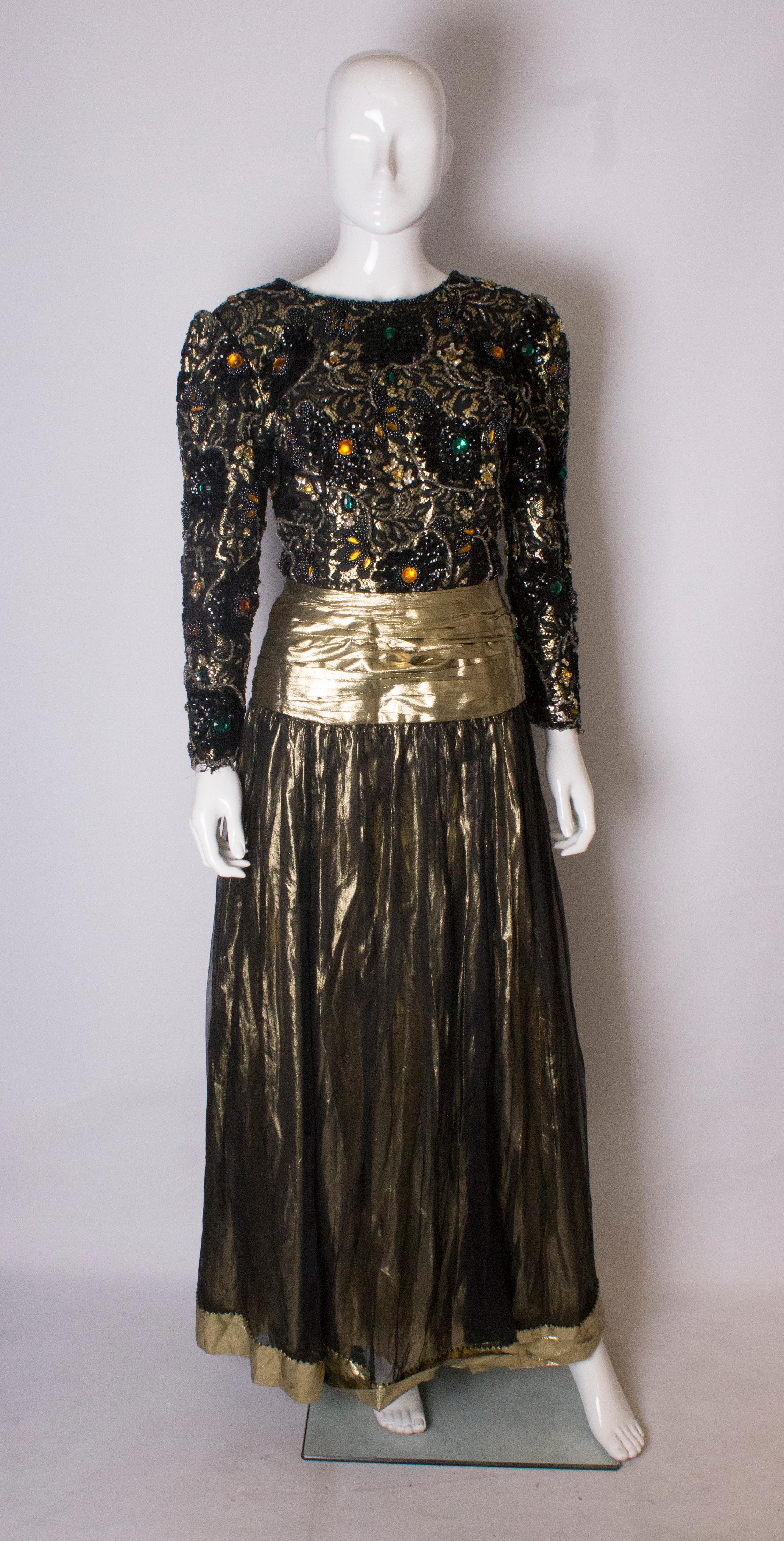A head turning evening gown from the 1980s. The dress has a heavily beaded top part with a deep v backline. The waist area is in a gold fabric , and the gathered skirt is a light black fabric over a gold lame underskirt.