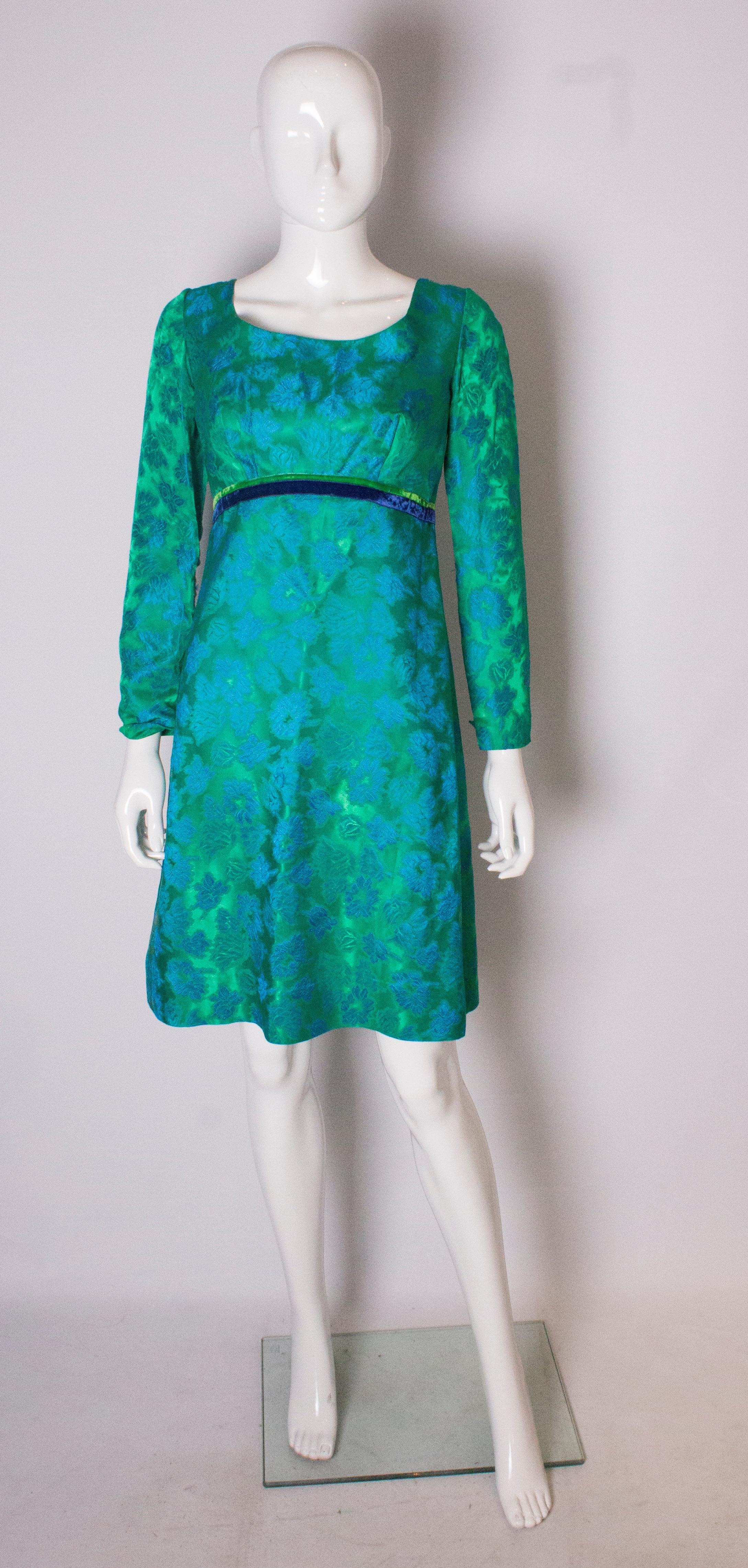 A chic cocktail dress from the 1960s. In a stunning green fabric with blue floral pattern, the dress has elbow length sleeves, blue and green ribbon detail under the bust , and a scoop neckline and backline. It is fully lined, with a 3''hem and