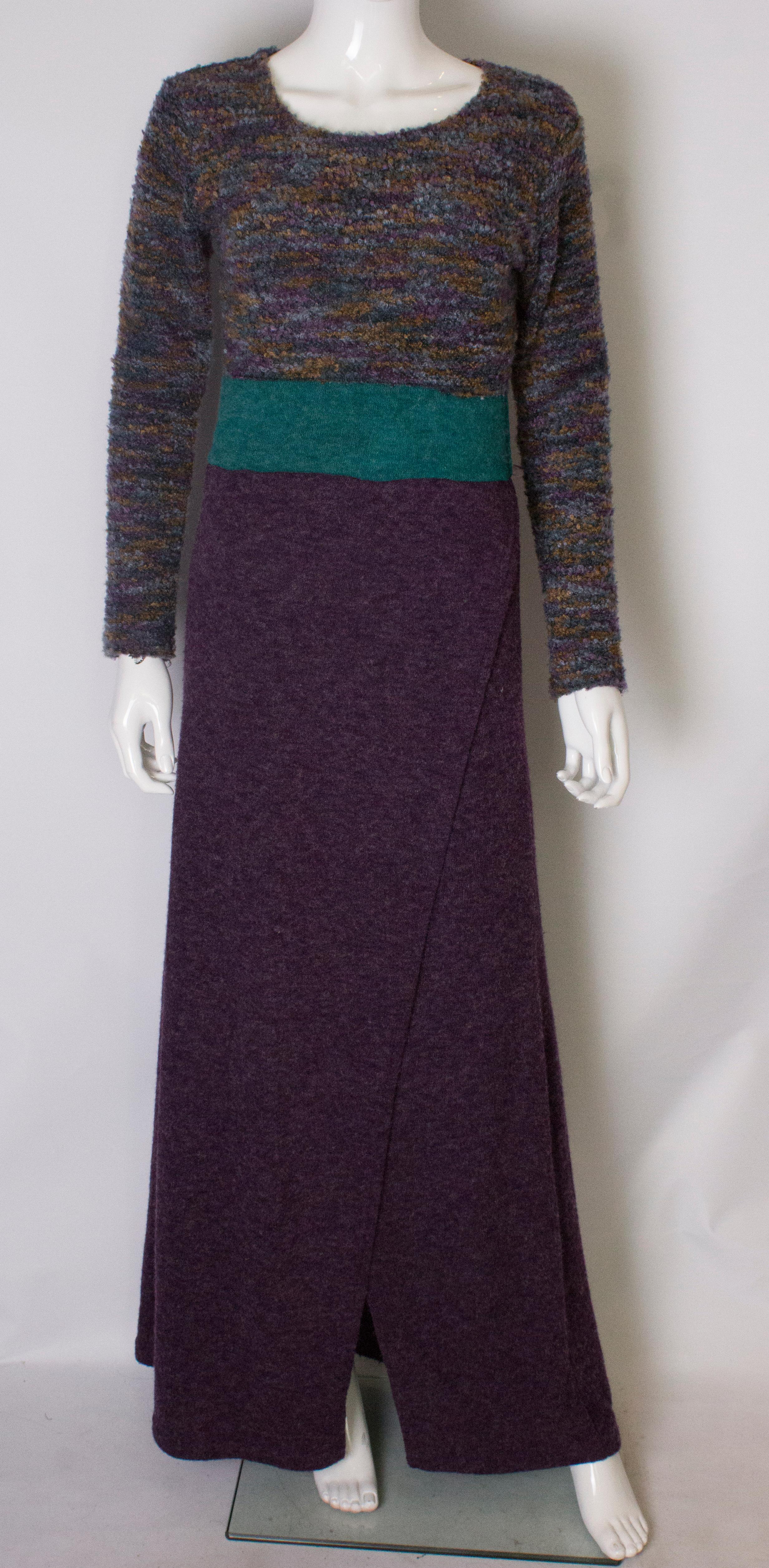 Une robe chic et chaleureuse dans un joli mélange de couleurs et de textures. La robe a une encolure dégagée, avec une partie supérieure violette et turquoise. Il y a une bande turquoise au milieu et une jupe violette avec une ouverture à