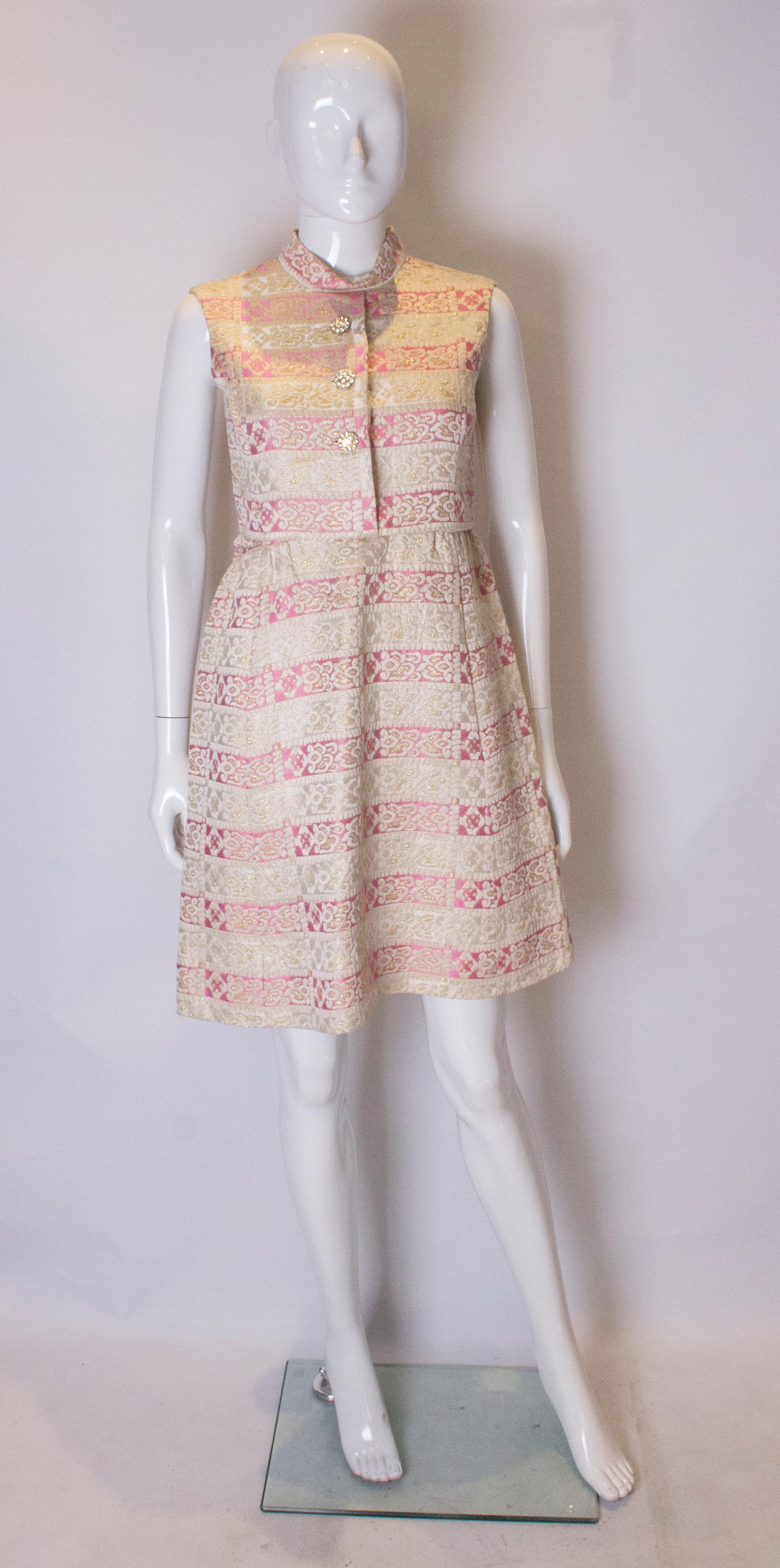 A chic cocktail dress from the 1960s.  The dress is in a pink and gold fabric, and is fully lined with a central back zip. It has a round neck, and decorative buttons at the front.  There are two pockets in the skirt part.