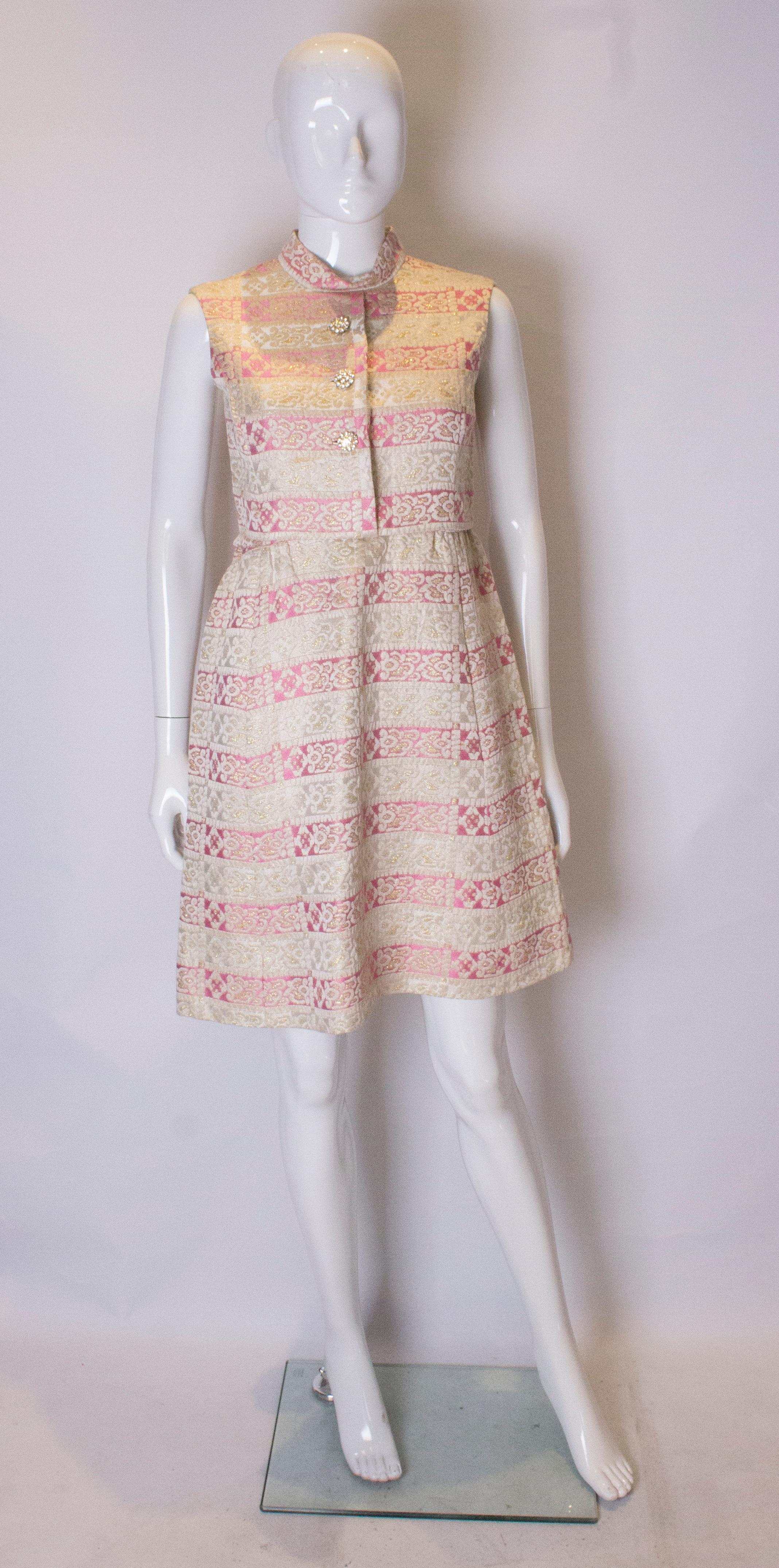 Ein schickes Cocktailkleid aus den 1960er Jahren.  Das Kleid ist in einem rosa-goldenen Stoff gehalten, vollständig gefüttert und hat einen zentralen Reißverschluss am Rücken. Es hat einen Rundhalsausschnitt und dekorative Knöpfe auf der
