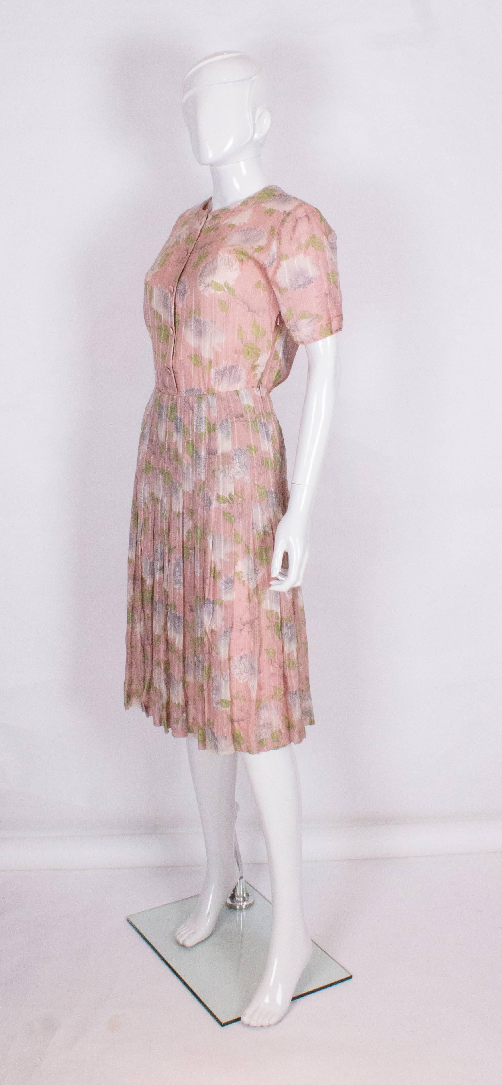 A stylish slk day dress.The dress has a pink background, with a green ,cream and grey floral design. It has a round neckline , with a 5 button opening at the front, and a side zip opening.There are 7'' sewn down pleats from the waist, giving a